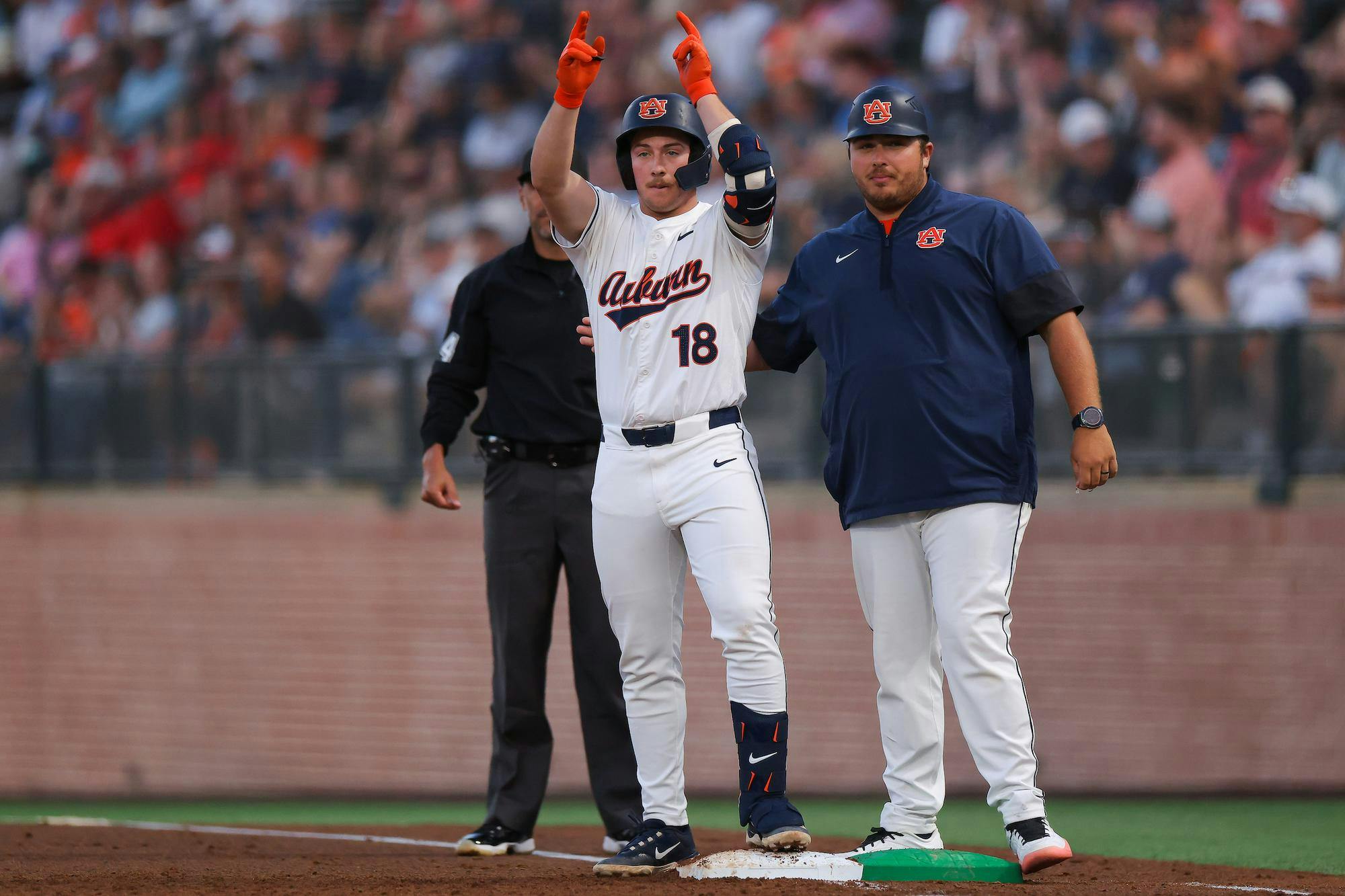 A baseball player in a white jersey with "Auburn" printed on it raises both arms in celebration, standing next to a coach.