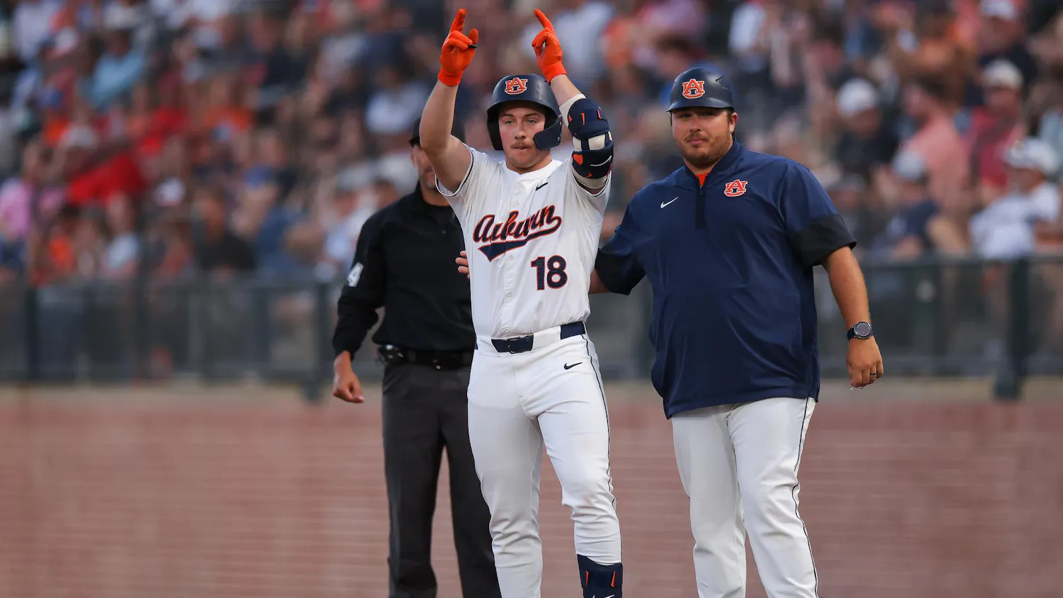 A baseball player in a white jersey with "Auburn" printed on it raises both arms in celebration, standing next to a coach.