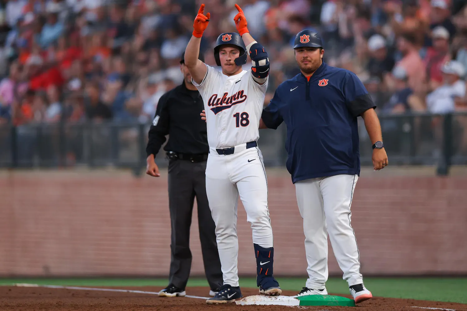 A baseball player in a white jersey with "Auburn" printed on it raises both arms in celebration, standing next to a coach.