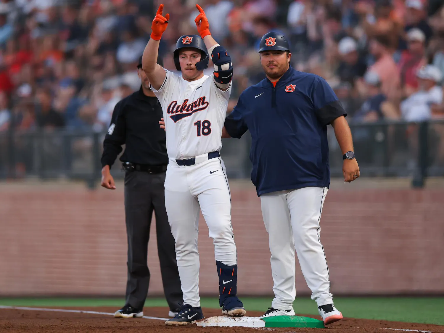A baseball player in a white jersey with "Auburn" printed on it raises both arms in celebration, standing next to a coach.