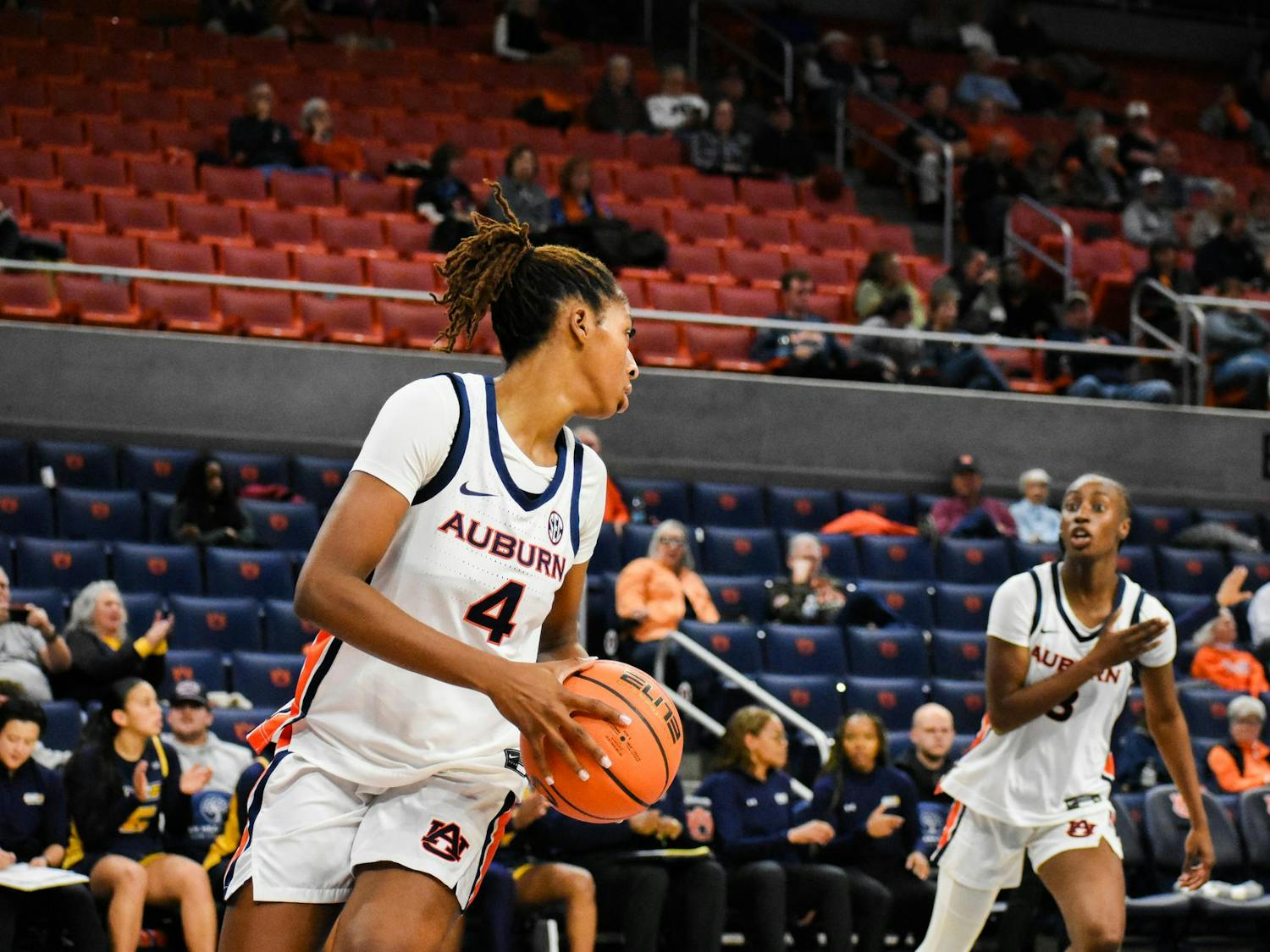 A basketball player in an Auburn uniform prepares to pass the ball while another teammate calls for it nearby, with spectators in the background.