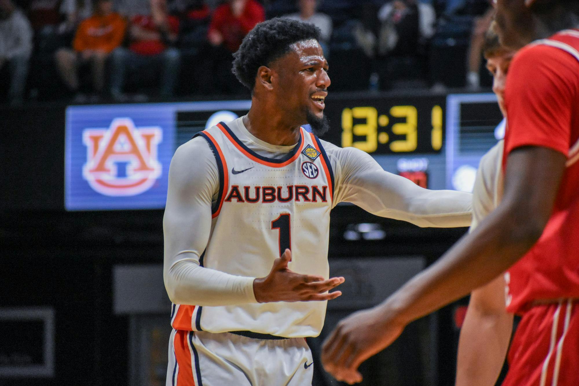 A basketball player in a white jersey with "Auburn" printed on it gestures passionately during a game, with a scoreboard in the background.