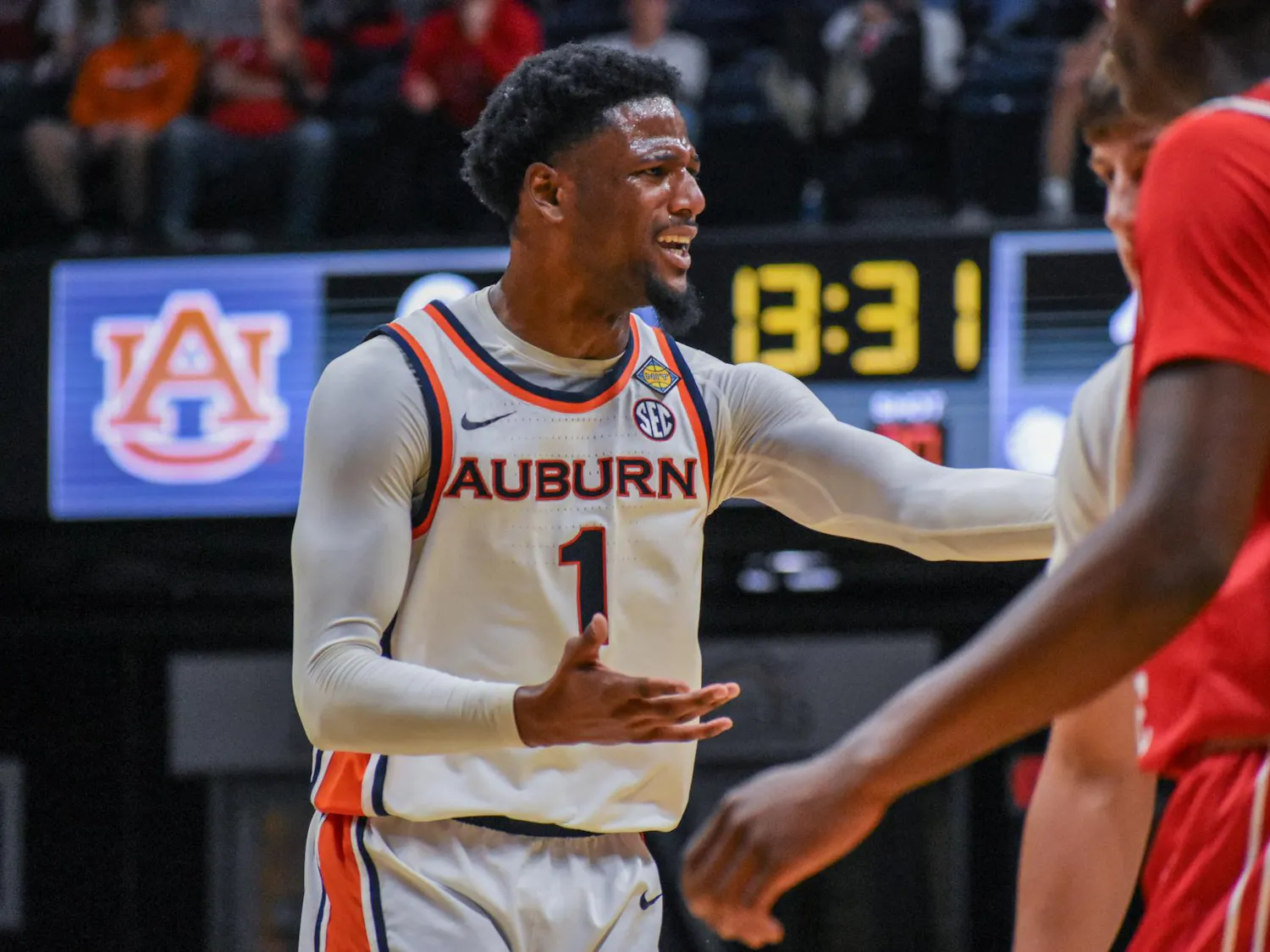 A basketball player in a white jersey with "Auburn" printed on it gestures passionately during a game, with a scoreboard in the background.