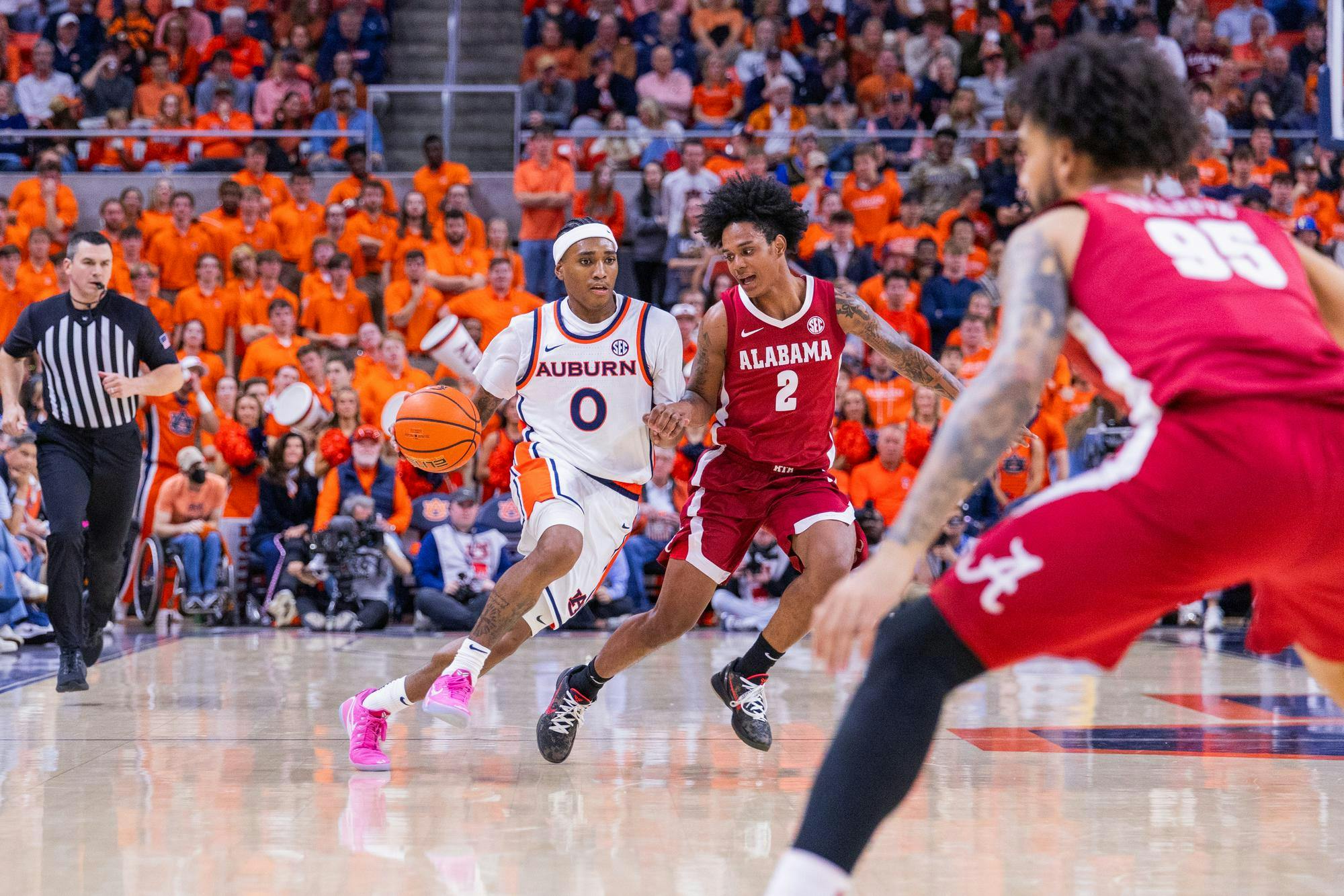A basketball player in a white Auburn uniform dribbles past an opponent in a red Alabama jersey, while a referee watches.
