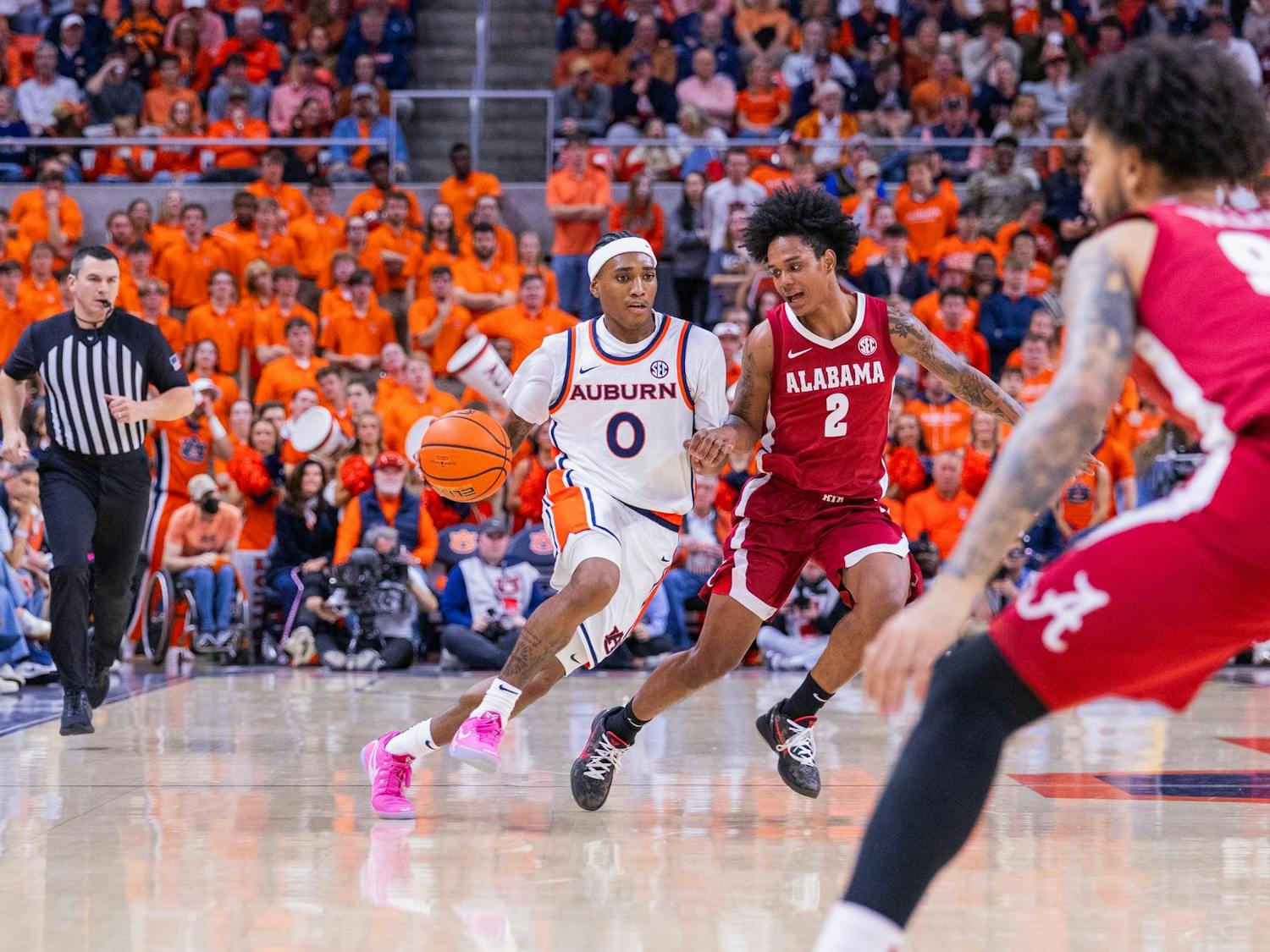 A basketball player in a white Auburn uniform dribbles past an opponent in a red Alabama jersey, while a referee watches.