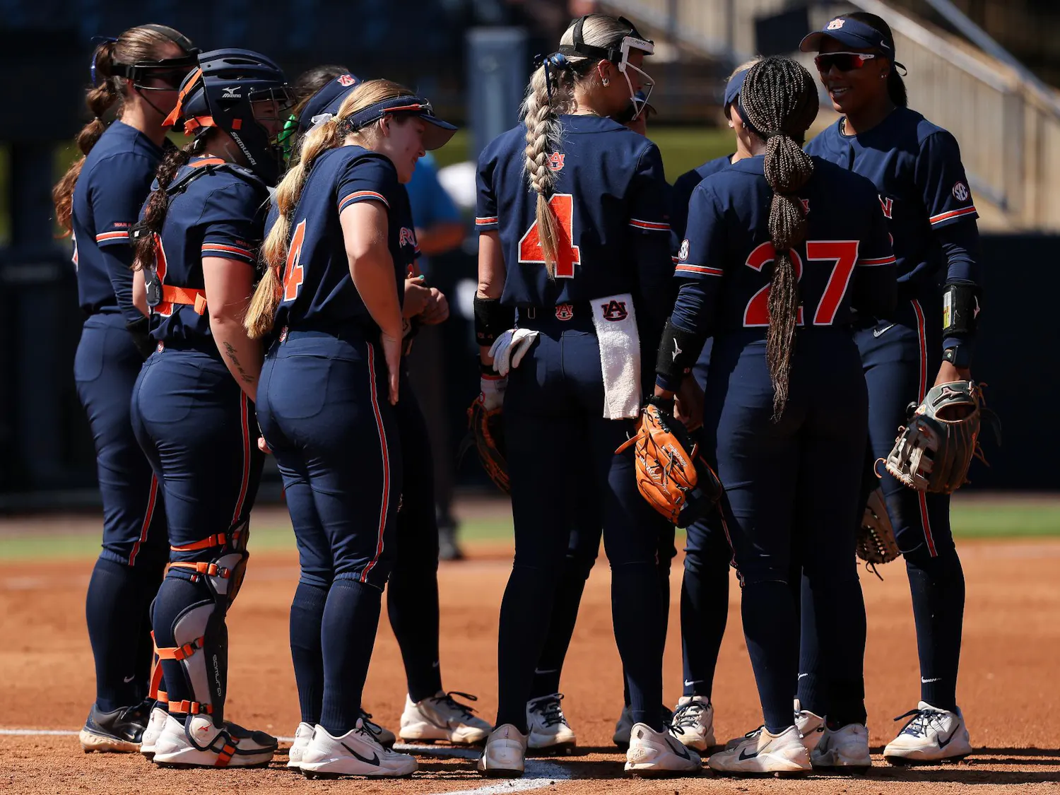 A group of eight female softball players in navy uniforms are gathered in a huddle on a dirt field.