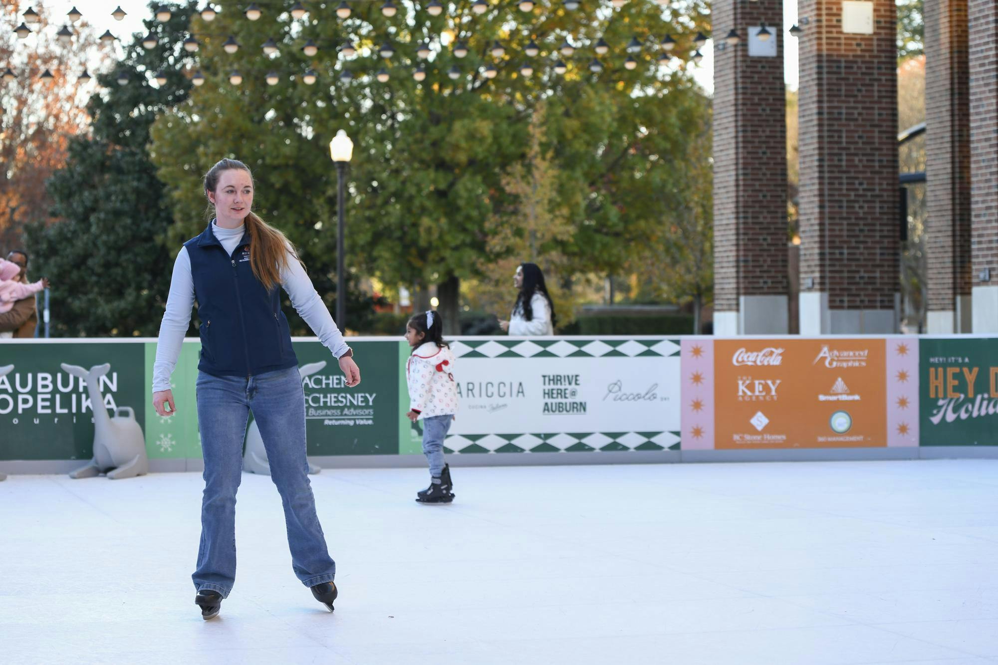 A woman ice skates on a rink, surrounded by trees and banners advertising various businesses, with children nearby.