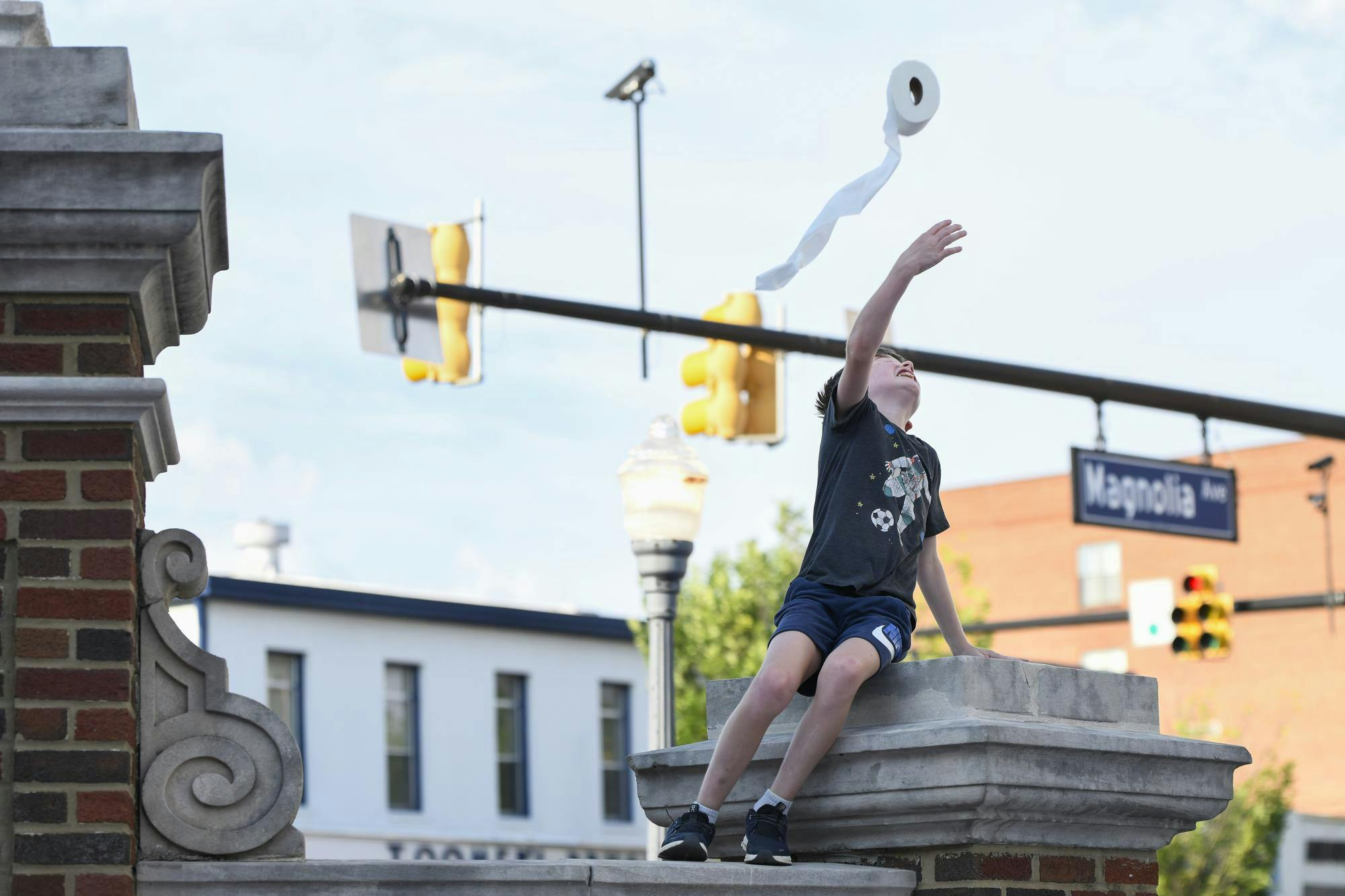 A boy sitting on a stone structure reaches up to throw a roll of toilet paper into the air.