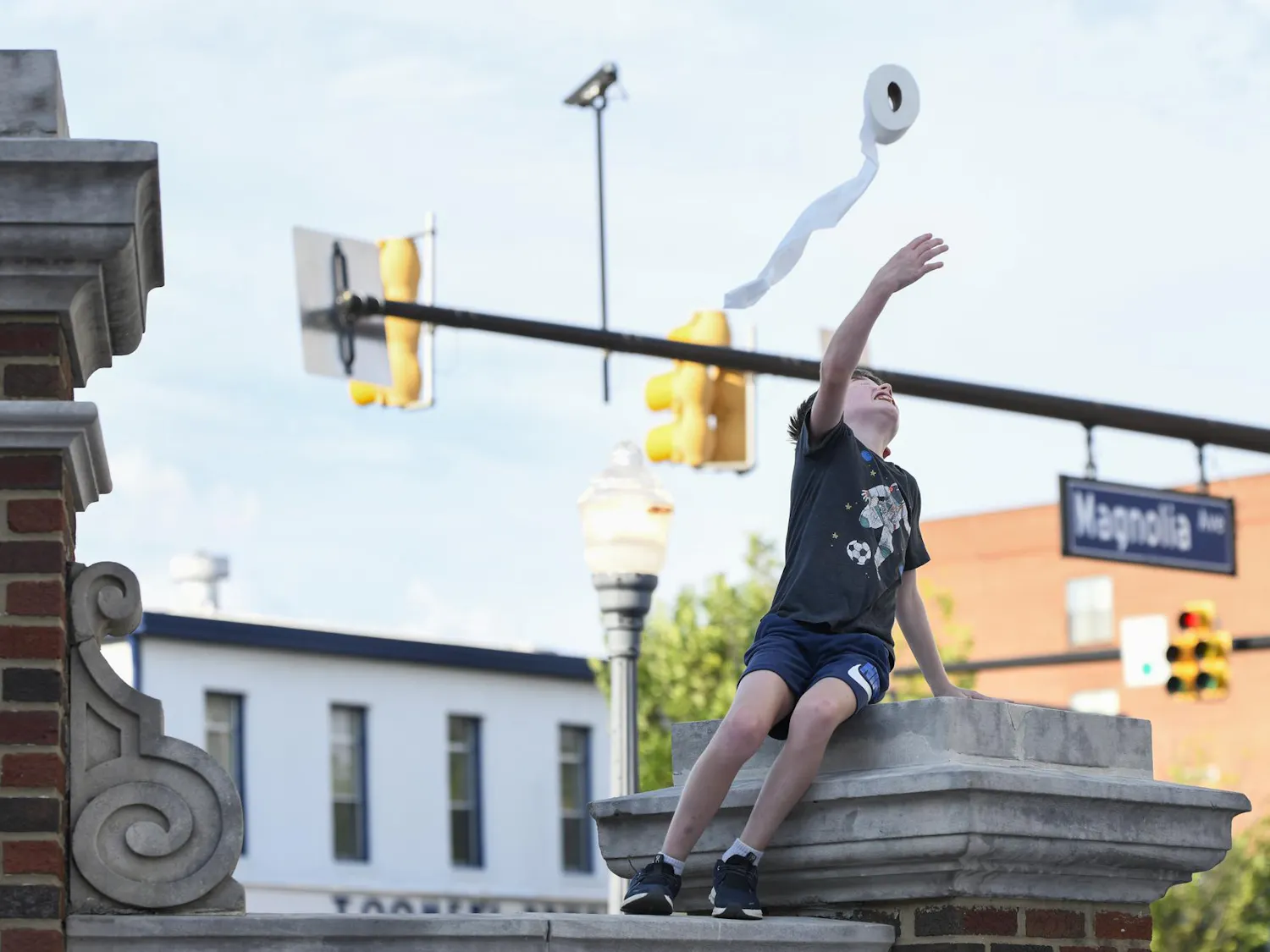A boy sitting on a stone structure reaches up to throw a roll of toilet paper into the air.