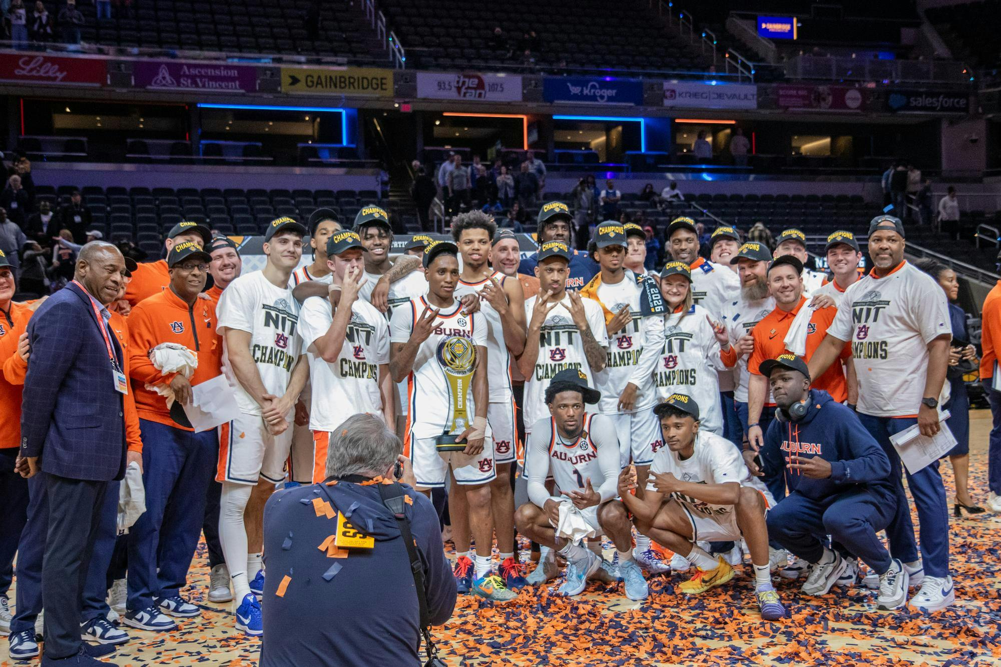 A group of athletes and coaches celebrate a championship victory, holding a trophy amidst confetti and wearing "champions" hats.