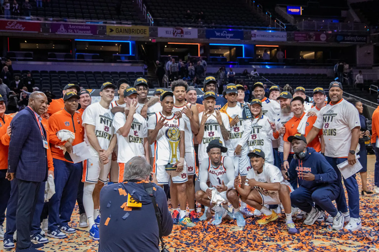 A group of athletes and coaches celebrate a championship victory, holding a trophy amidst confetti and wearing "champions" hats.