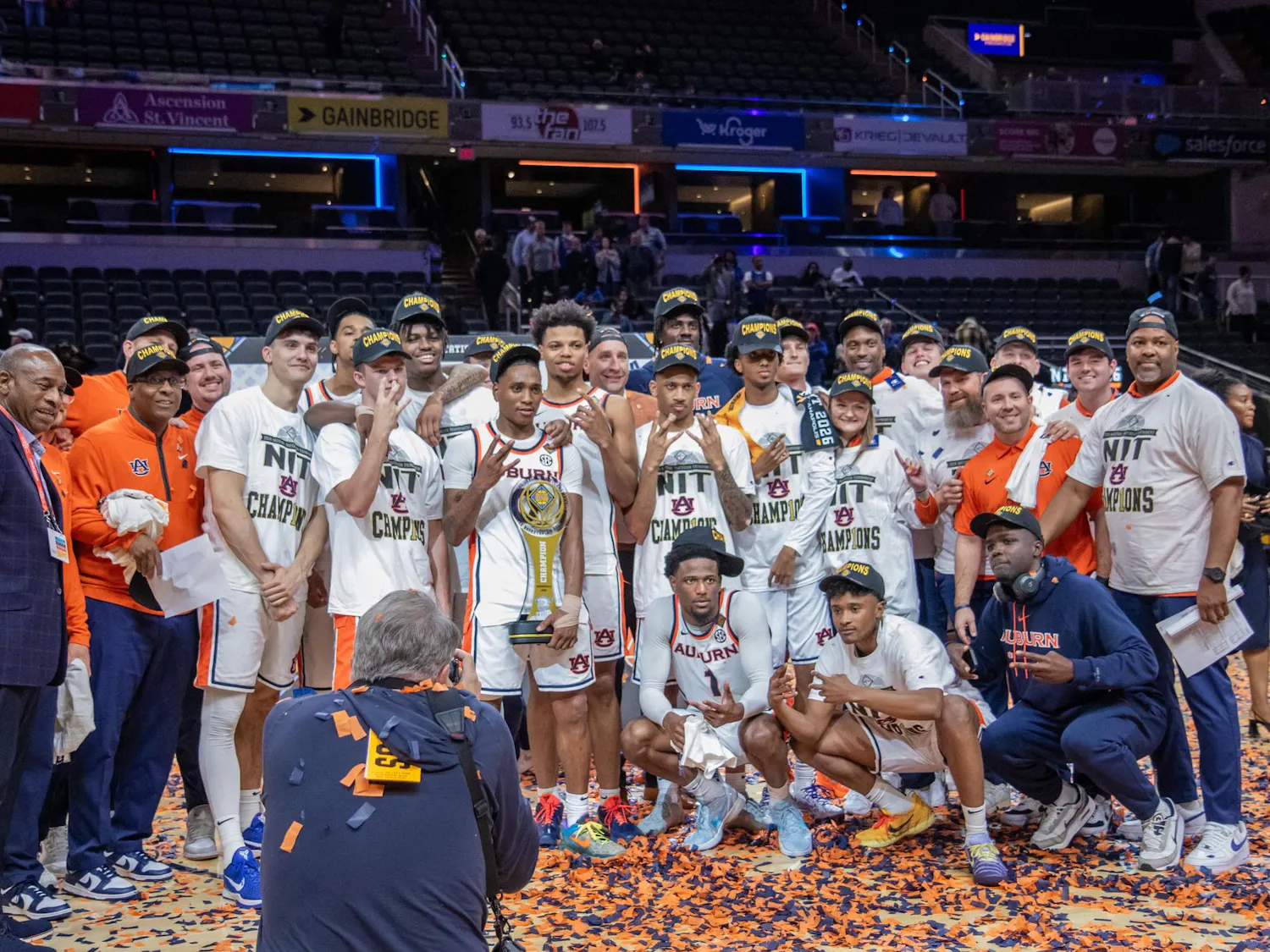 A group of athletes and coaches celebrate a championship victory, holding a trophy amidst confetti and wearing "champions" hats.