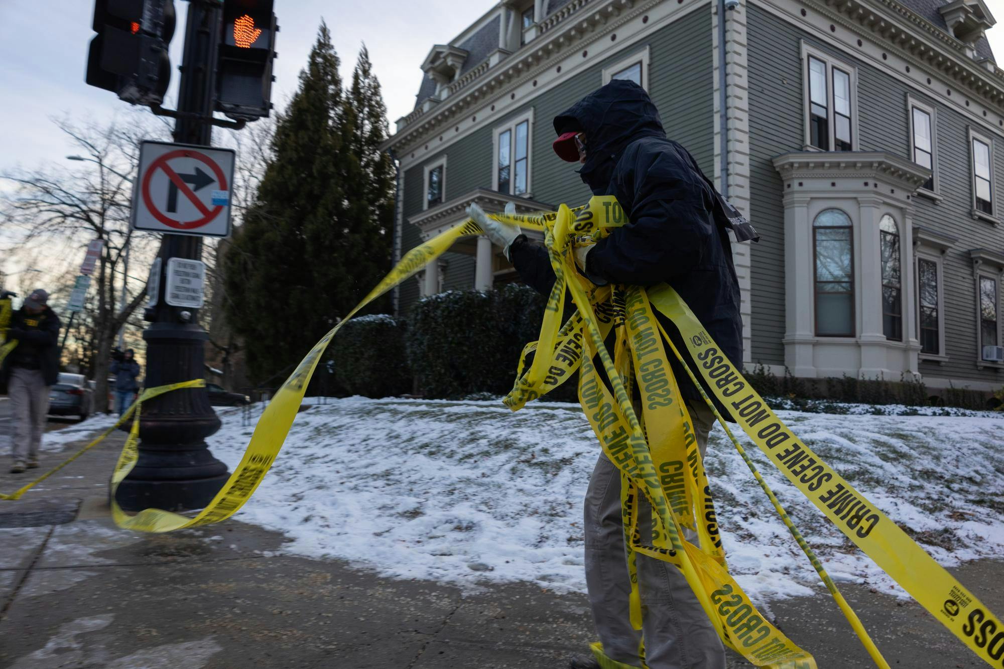 A person in a black coat, seen wearing FBI gear, walks by a street corner carrying caution tape that appears to have been ripped down. 