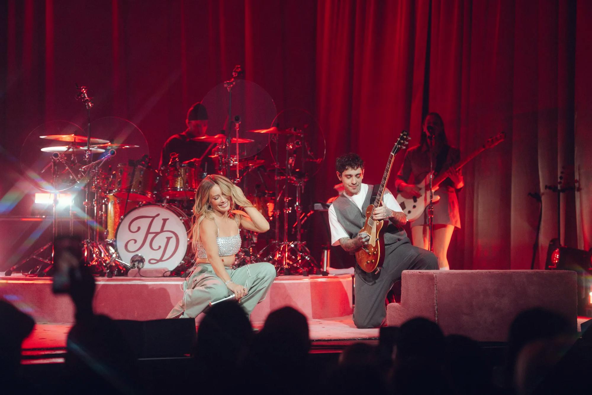 Photo of Hilary Duff in a silver top and gray pants performing on a red stage with large curtains. A band surrounds her.  
