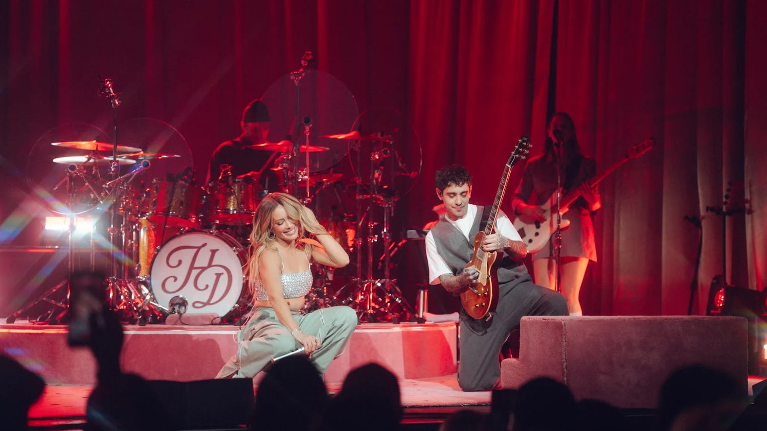 Photo of Hilary Duff in a silver top and gray pants performing on a red stage with large curtains. A band surrounds her.