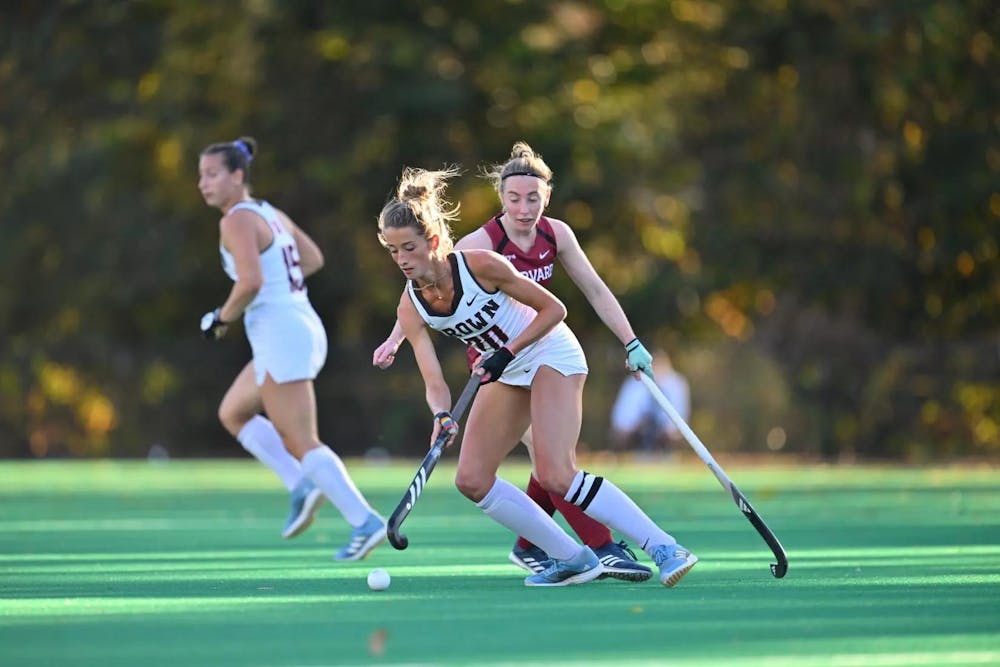 Two field hockey players battle for a puck. The Brown University photo is wearing a white jersey with the ball running away from the Harvard player in red.