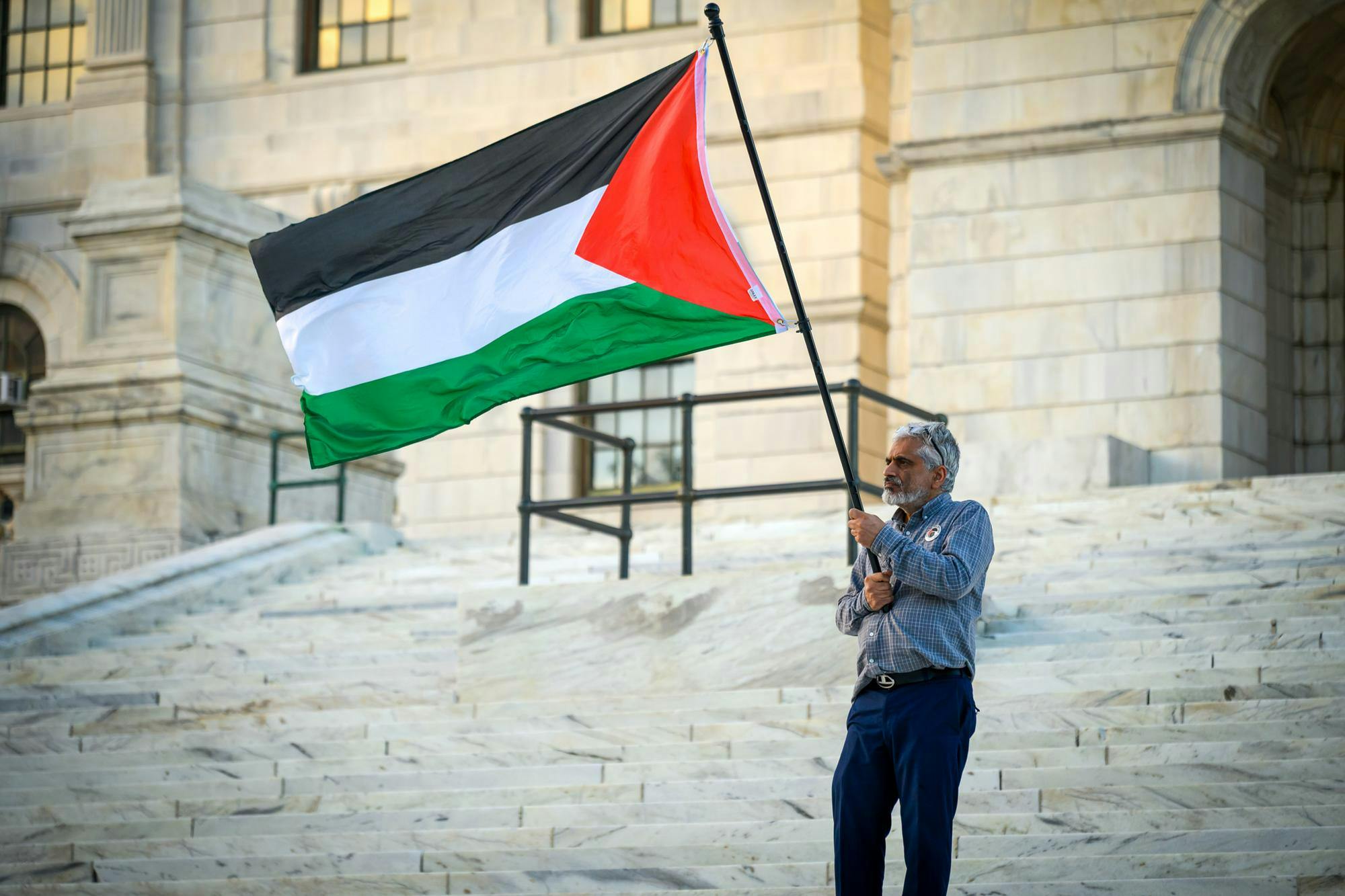 A man holds a large flag of Palestine in front of marble steps.