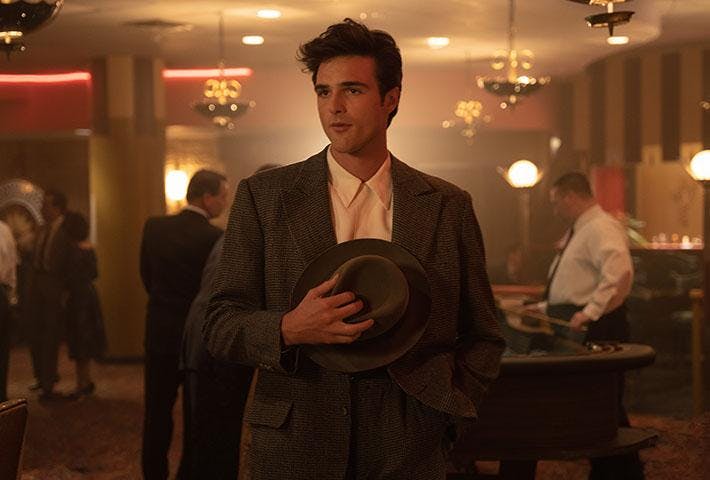 A man in a casino hall stands in a brown suit holding a brown hat to his chest.