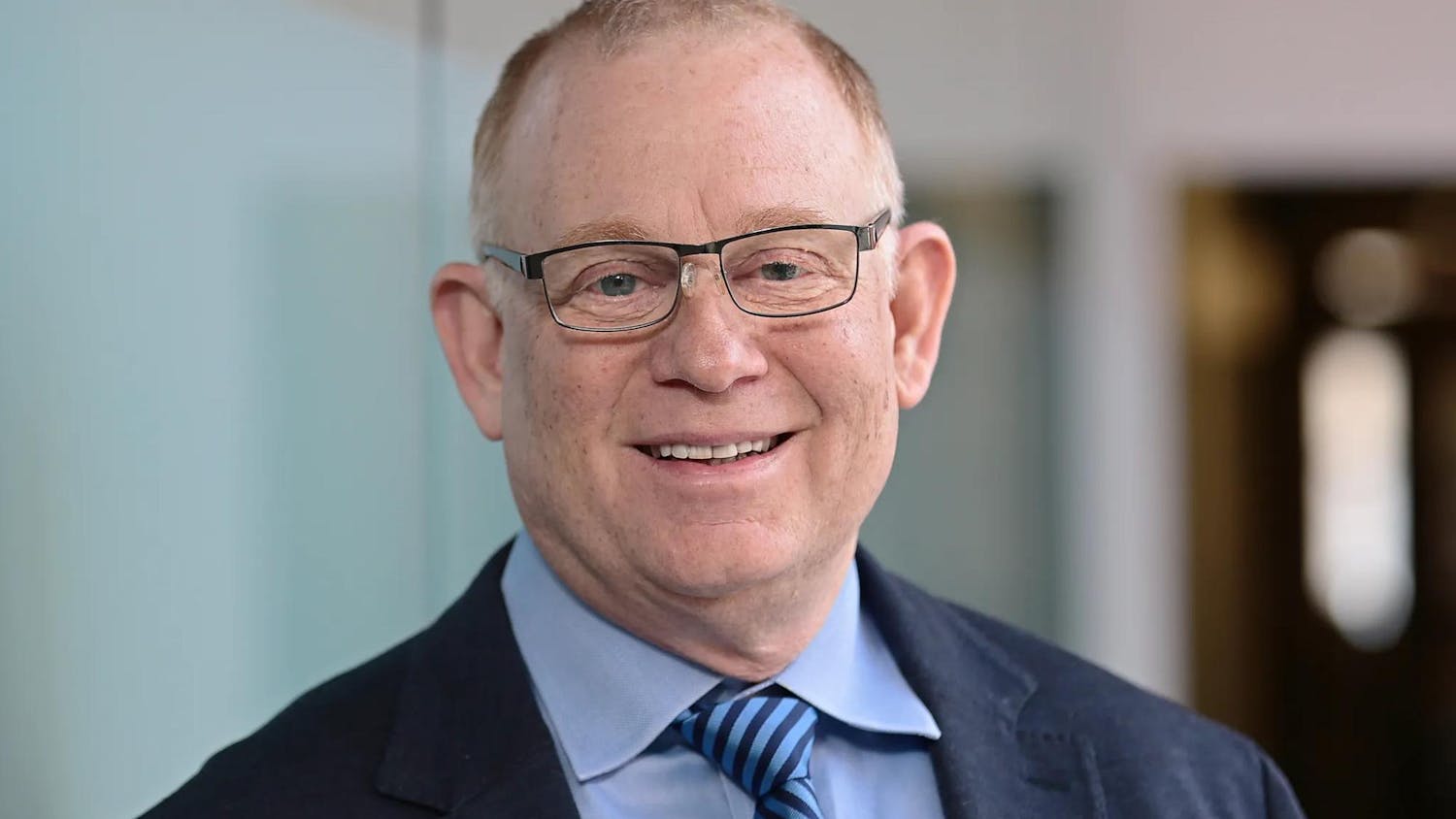 Up close portrait shot of Michael Satlow, who wears glasses and a blue suit, smiling against a blurred background.