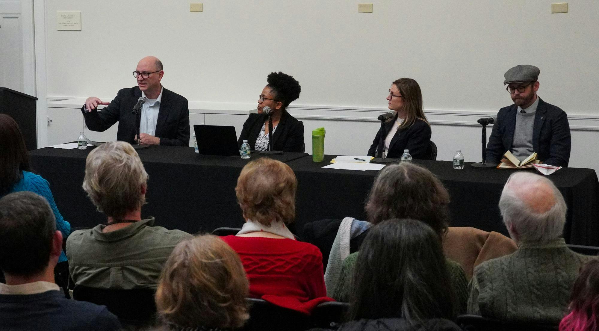 Four individuals wearing business attire sit at a long table covered in a black tablecloth in front of an audience. 