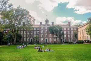 Picture of the University Hall on a sunny day with surrounding trees and multiple groups of students scattered around the vicinity.