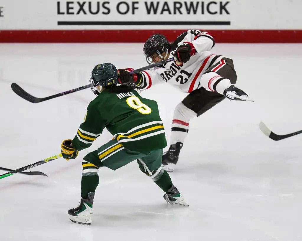 A photo of a hockey player hitting a puck with their hockey stick skating next to another player. 