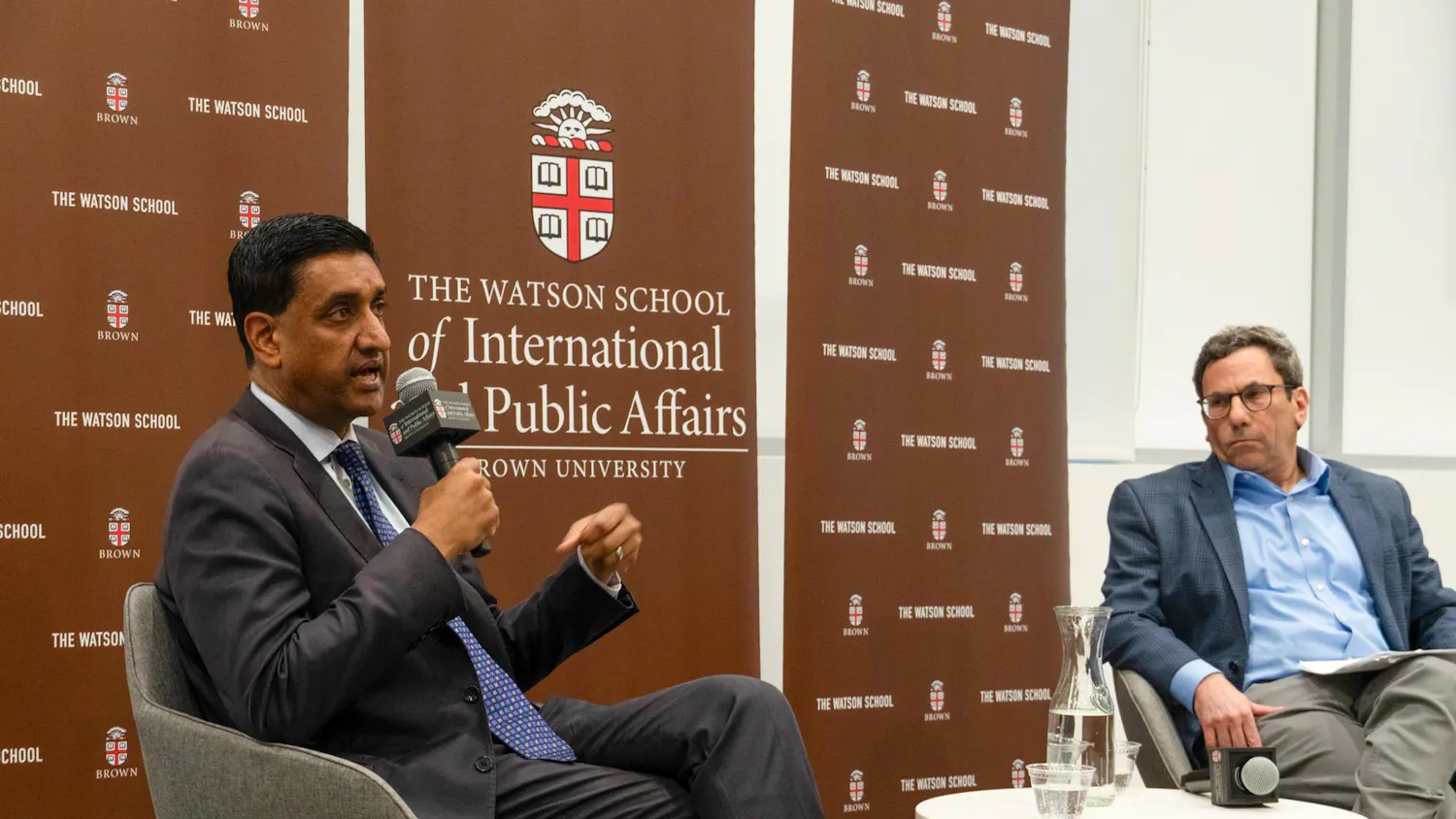Ro Khanna, in a grey suit, sits in a chair next to moderator Eric Patashnik, director of the Taubman Center. The two are in front of a brown backdrop.