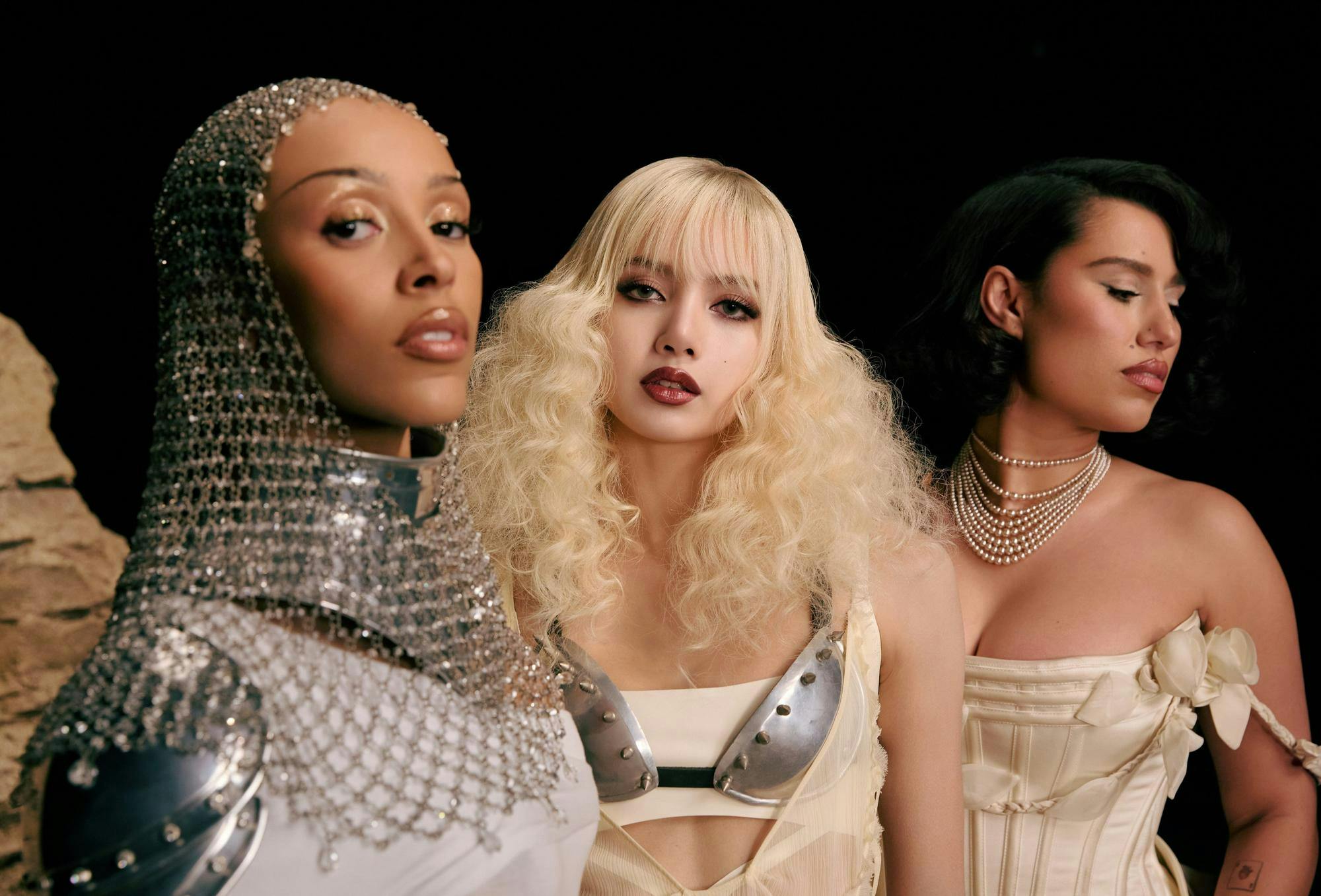 A picture of Doja Cat, Lisa and Raye on the set of the "Born Again" music video. Doja Cat is in the front wearing chain mail on her head, Lisa is in the middle with a metal breastplate and blonde hair and Raye is in the back with a pearl necklace and white dress.