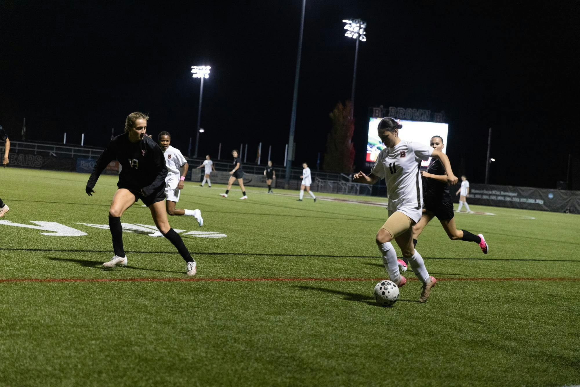 Photo of Claire Silverman ’27 in white shirt with a brown stripe running down the side and the number 11 on the front and white shorts. She is kicking a soccer ball in front of two players from Harvard.