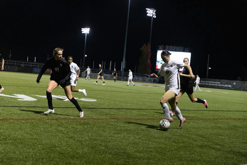 Photo of Claire Silverman ’27 in white shirt with a brown stripe running down the side and the number 11 on the front and white shorts. She is kicking a soccer ball in front of two players from Harvard.