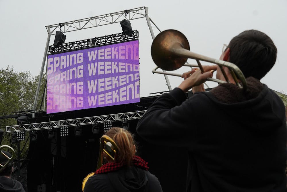 A photo of a person playing a trombone while facing toward a stage on the Main Green at Brown University. There is a screen that displays "Spring Weekend" in a funky font several times.