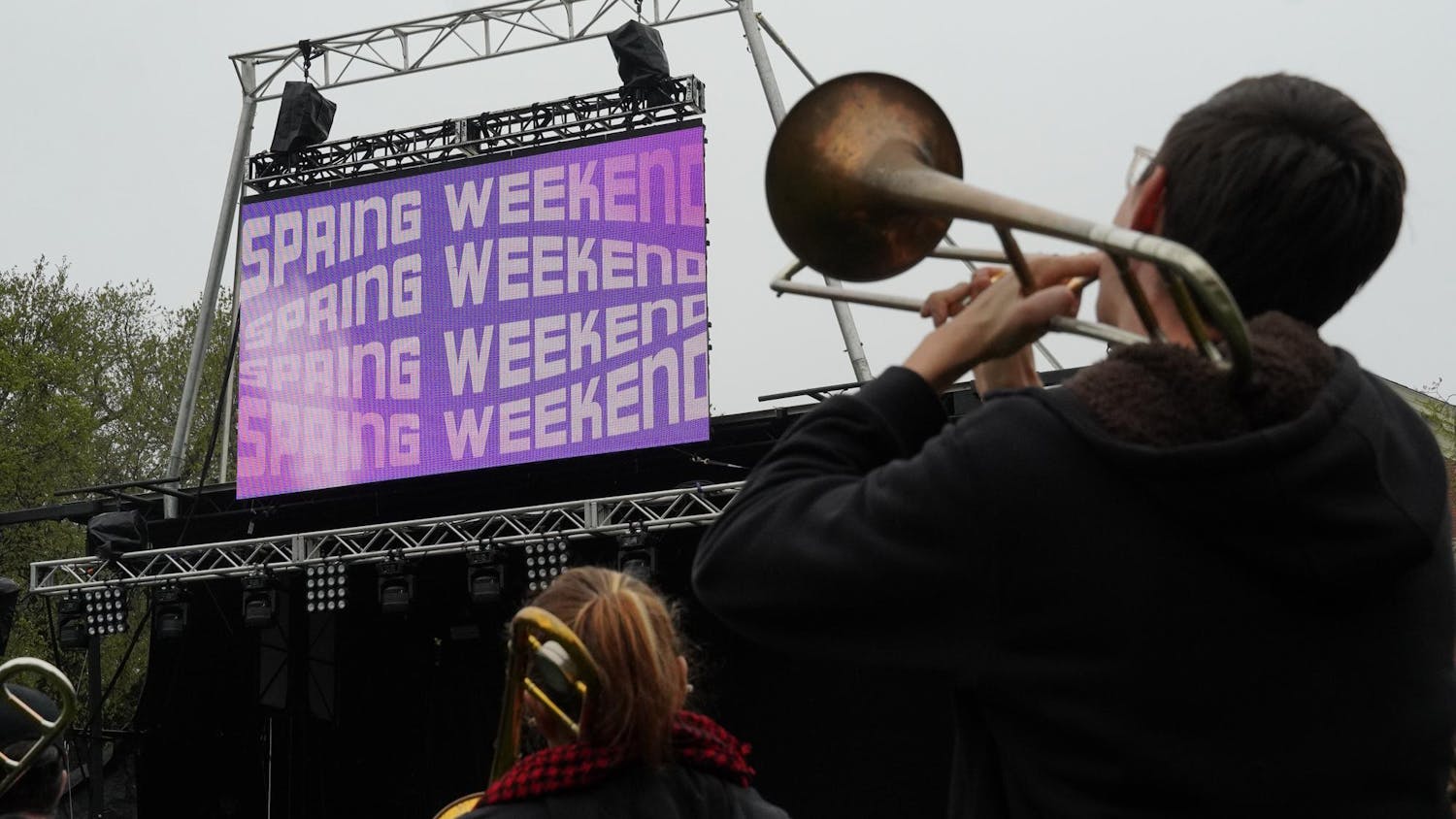 A photo of a person playing a trombone while facing toward a stage on the Main Green at Brown University. There is a screen that displays "Spring Weekend" in a funky font several times.