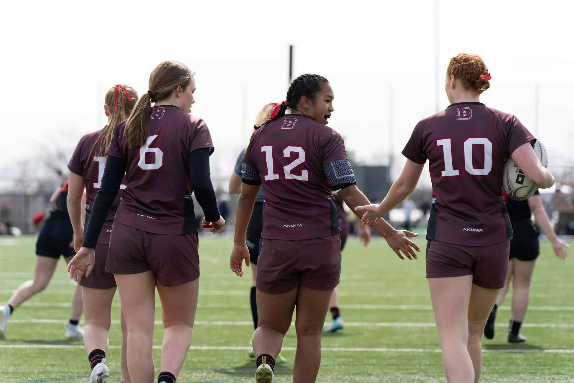 Three Brown women's rugby players in uniform with their backs to the camera are pictured walking on the field, with the player on the right holding the ball in her right hand. 
