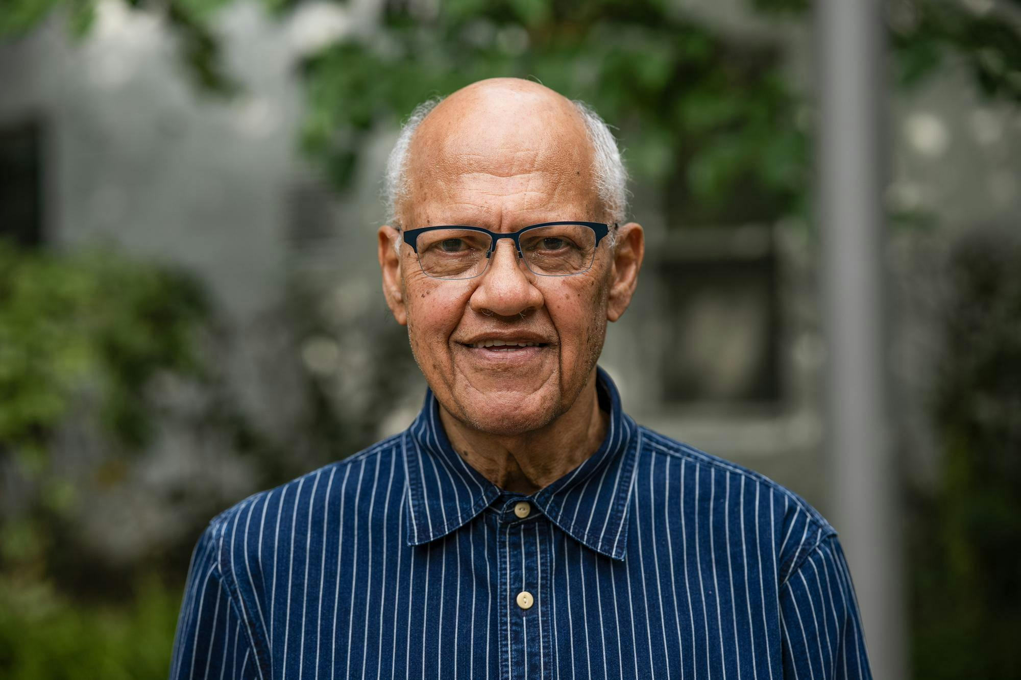 Bust photo of Dr. Wilmot James wearing a dark blue shirt with thin white stripes standing in front of a blurred background of green leaves and a gray building.