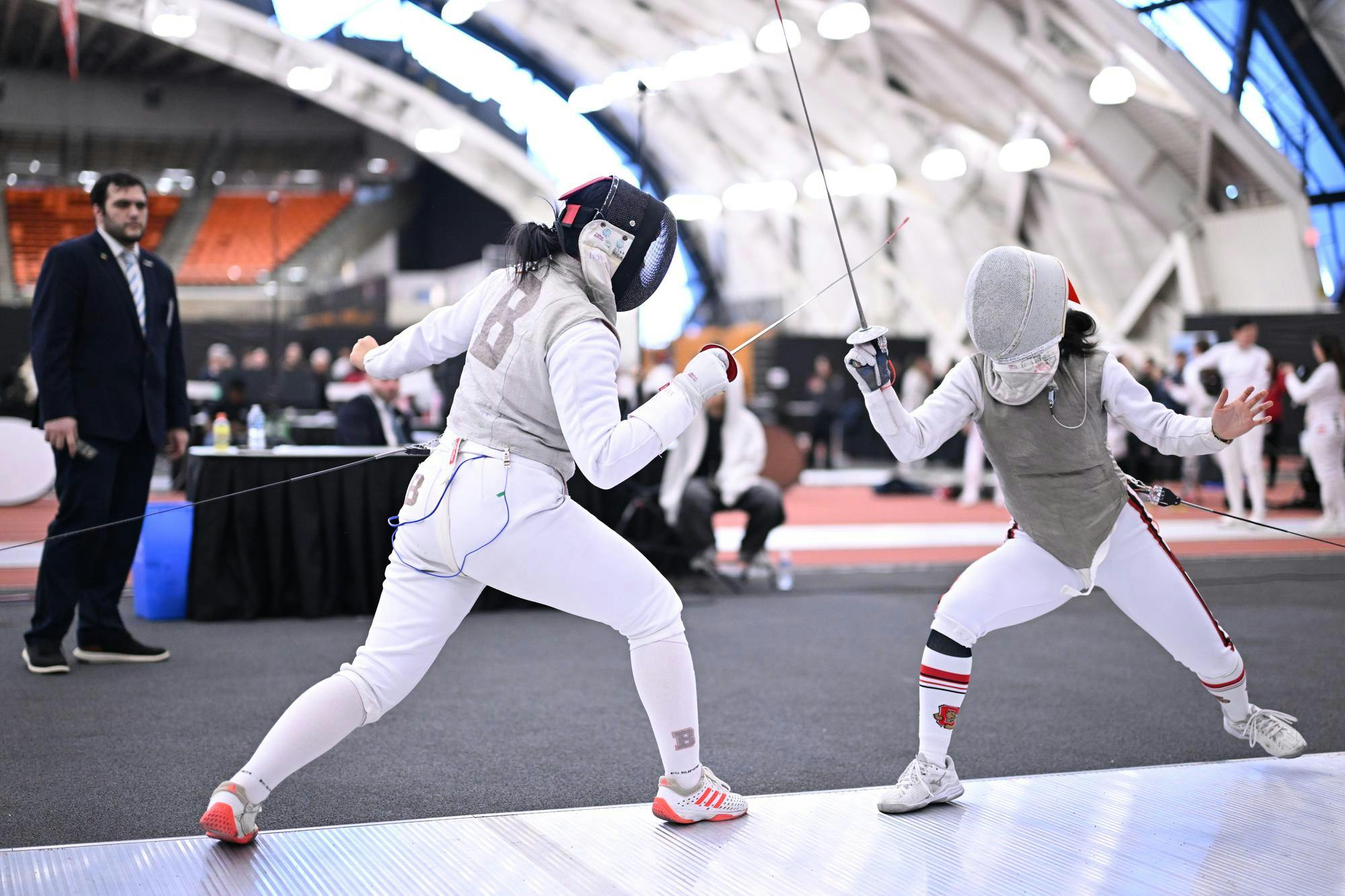 Photo of two fencers fencing, the fencer on the left has the Brown University logo on her back, looking down towards the fencer on the right.

