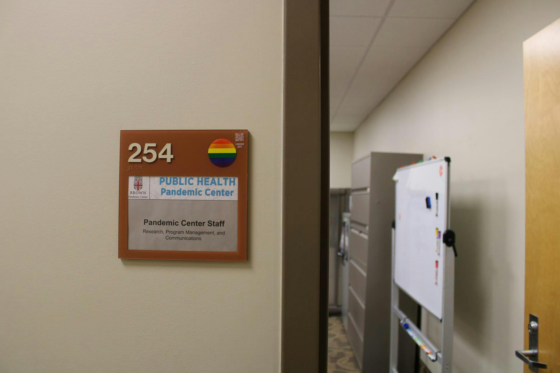 Photo of a sign with the number “254” in the left corner and a rainbow sticker on the right, with the Brown University insignia below, and the words “Public Health Pandemic Center.” The sign also displays the words “Pandemic Center Staff Research Program Management and Communications.” The sign is left of a door looking into an office with a whiteboard and filing cabinets.