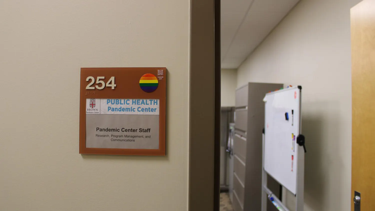 Photo of a sign with the number “254” in the left corner and a rainbow sticker on the right, with the Brown University insignia below, and the words “Public Health Pandemic Center.” The sign also displays the words “Pandemic Center Staff Research Program Management and Communications.” The sign is left of a door looking into an office with a whiteboard and filing cabinets.