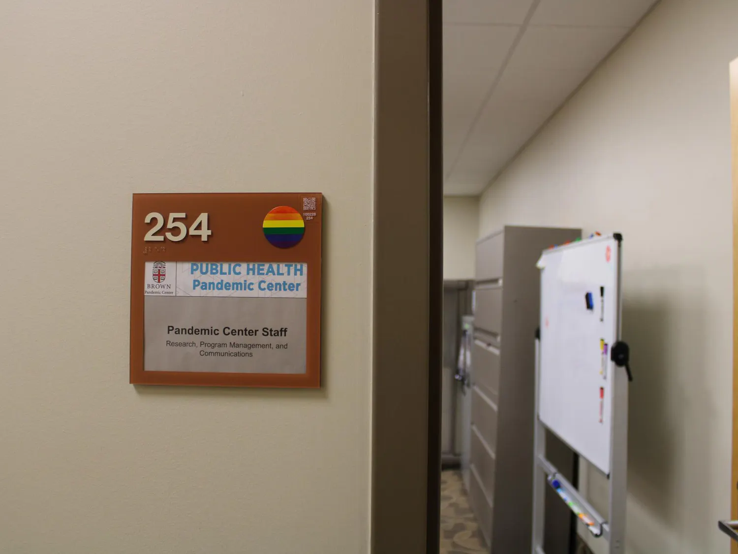 Photo of a sign with the number “254” in the left corner and a rainbow sticker on the right, with the Brown University insignia below, and the words “Public Health Pandemic Center.” The sign also displays the words “Pandemic Center Staff Research Program Management and Communications.” The sign is left of a door looking into an office with a whiteboard and filing cabinets.