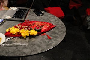 Photo of a red fan on top of a table with red pockets and a little green box with a red square and black lettering on top. A branch of yellow flowers sits on top of it. A computer is behind the setup with a phone and an iPad showing a student’s work.

