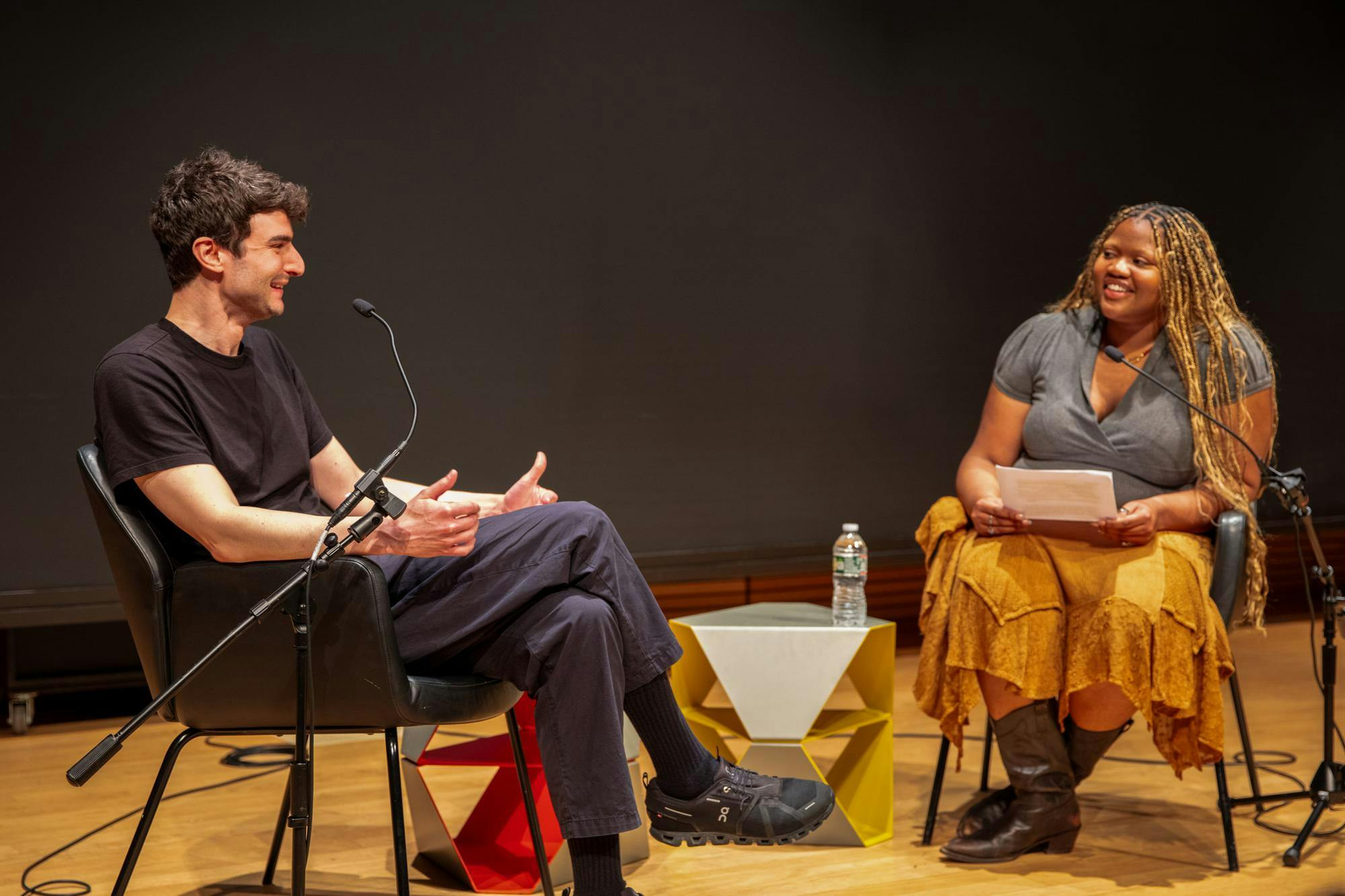 The picture shows alum and filmmaker Justin Kuritzkes talking with Herald-staffer and moderator Kendall Ricks. Kuritzkes is wearing a black shirt, dark blue pants and is smiling, while Ricks is wearing a yellow skirt and gray blouse. 