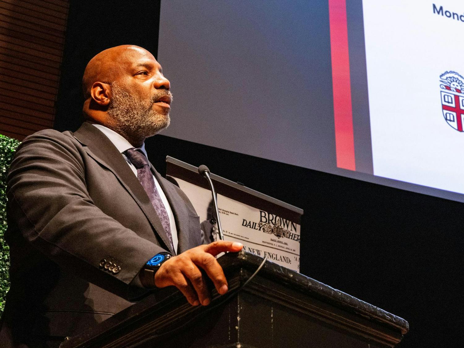 A man in a suit standing at a lectern with his mouth slightly open.