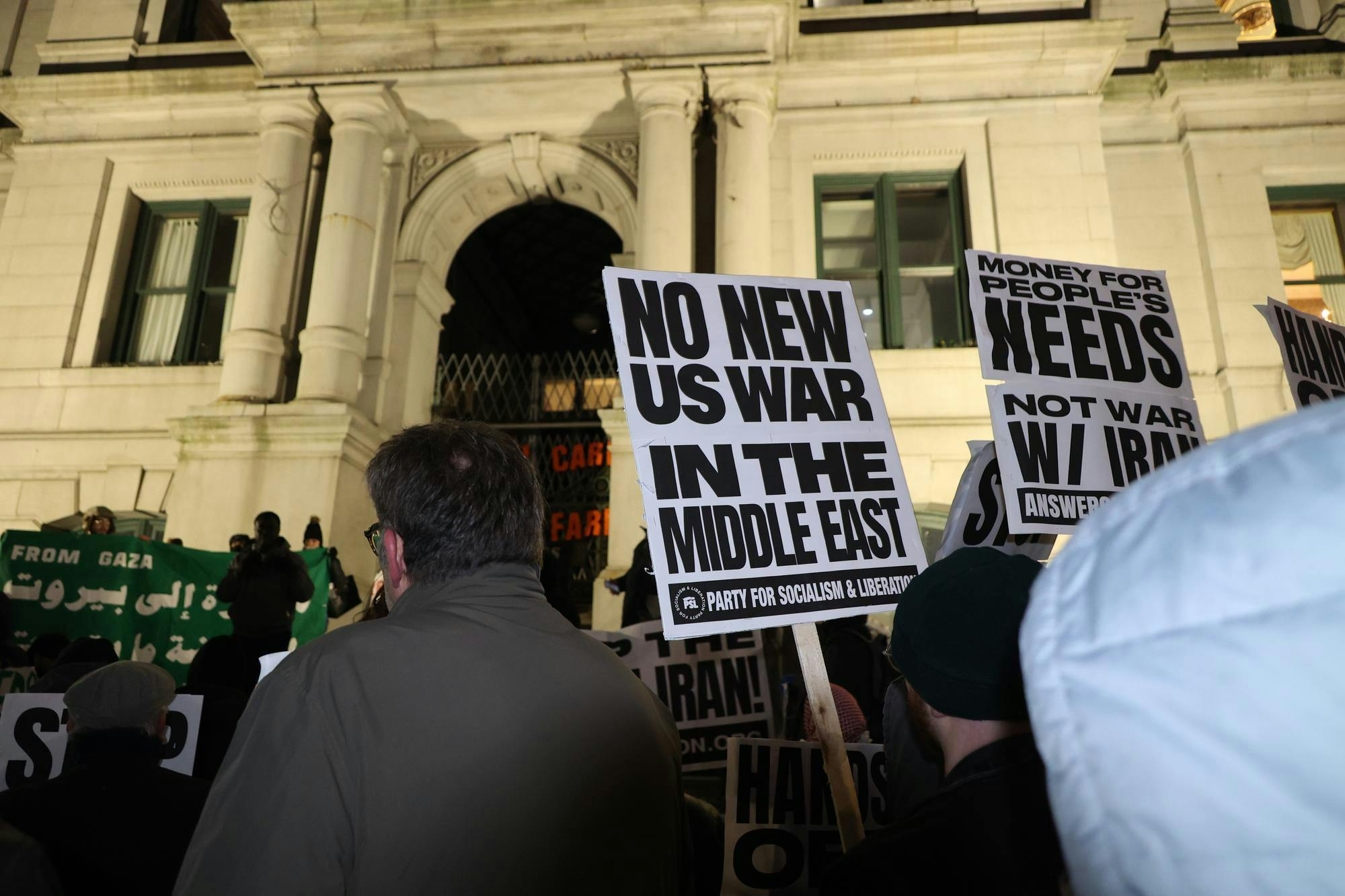 A photo showing protesters at night holding signs that read “No New U.S. War in the Middle East” in front of a white building with pillars. 