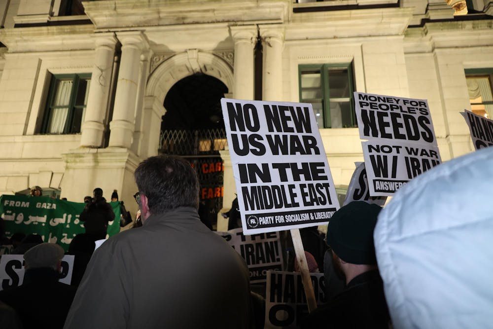 A photo showing protesters at night holding signs that read “No New U.S. War in the Middle East” in front of a white building with pillars. 