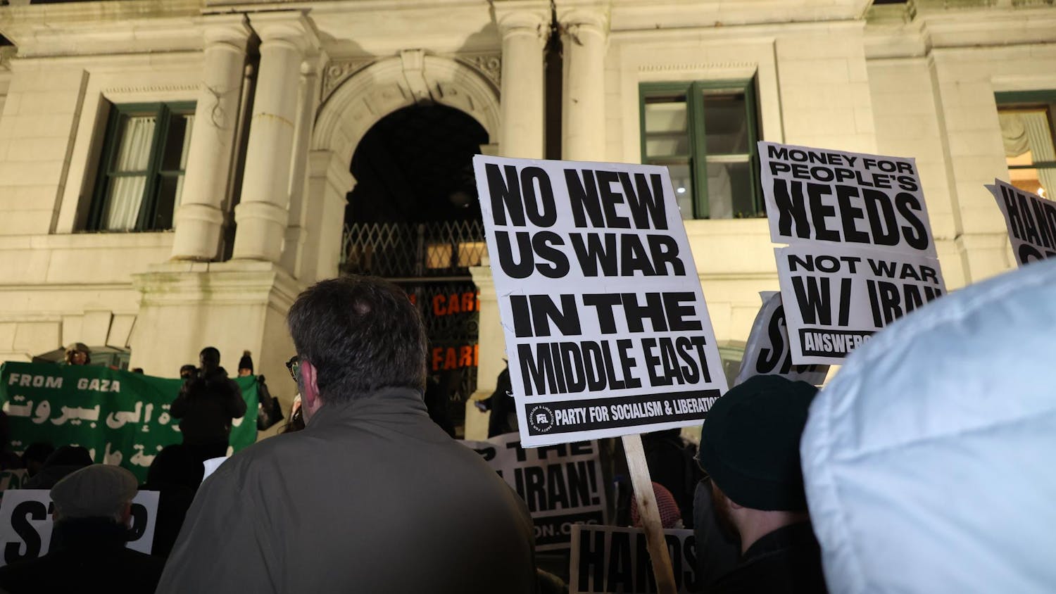 A photo showing protesters at night holding signs that read “No New U.S. War in the Middle East” in front of a white building with pillars.