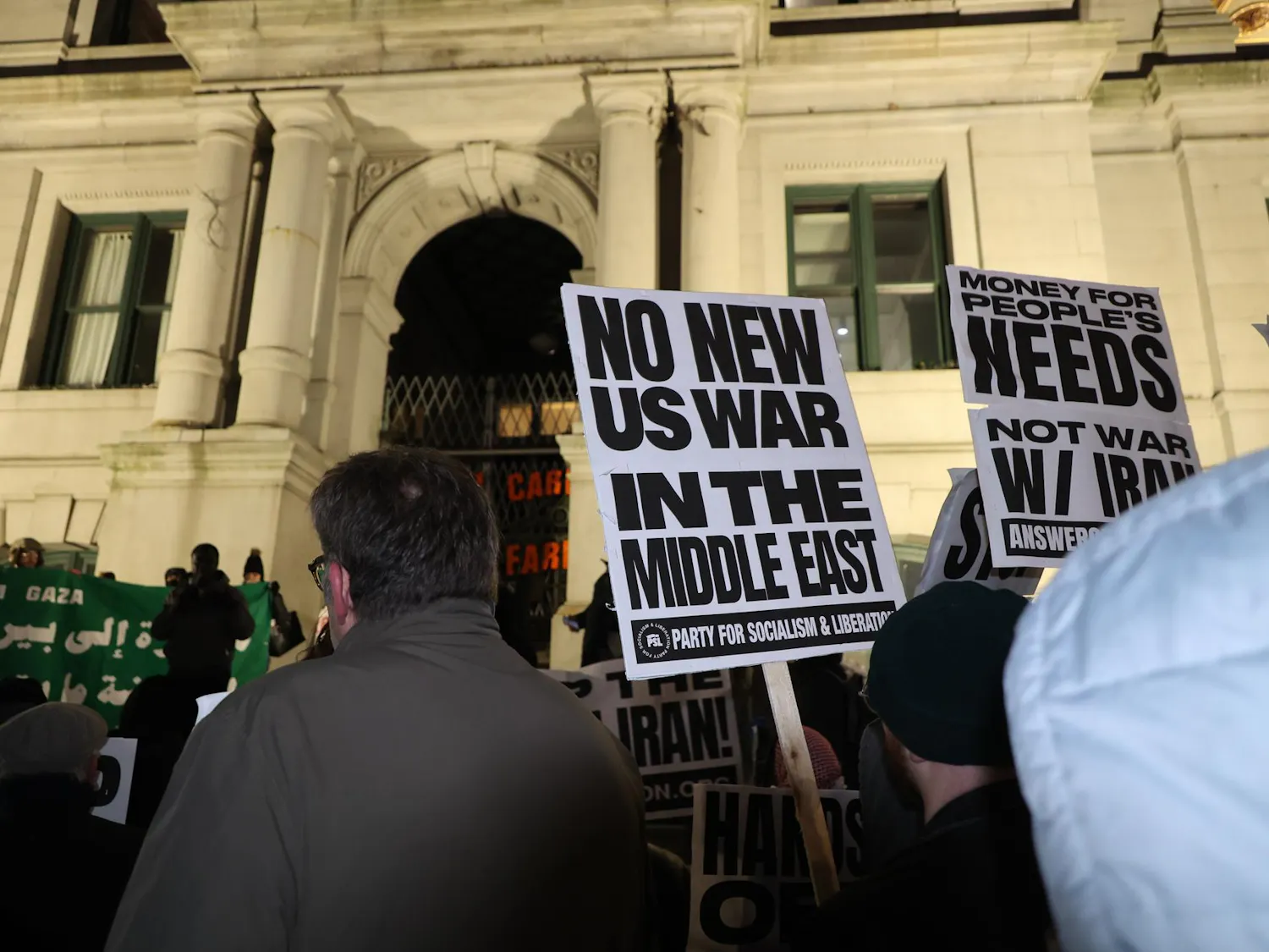 A photo showing protesters at night holding signs that read “No New U.S. War in the Middle East” in front of a white building with pillars.