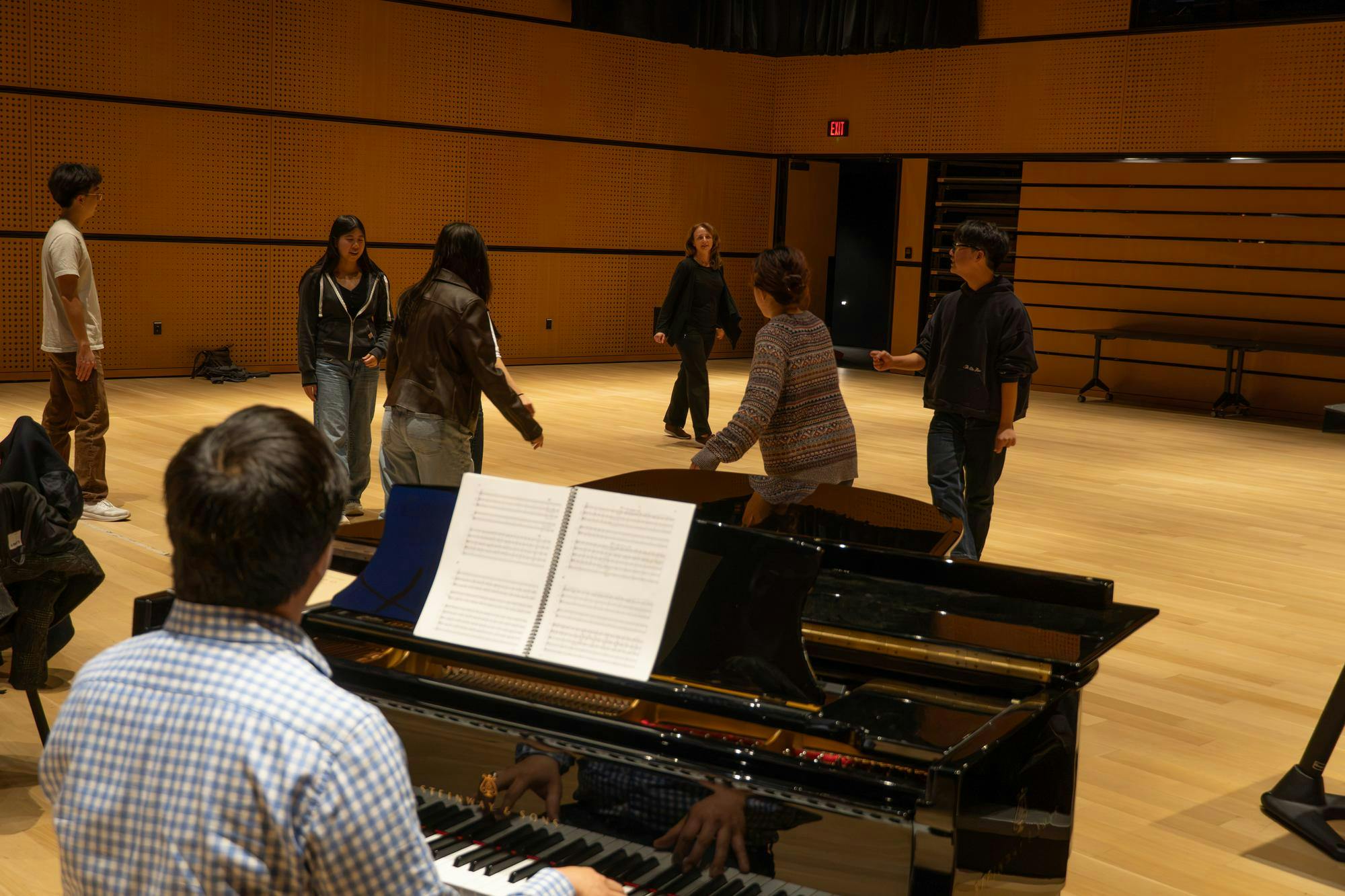 Photo of a man playing a piano with sheet music on the music stand. A group of people behind the piano are learning a dance from a professor.