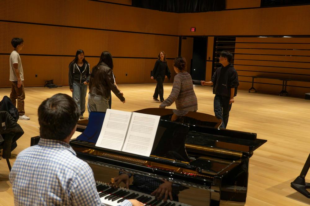 Photo of a man playing a piano with sheet music on the music stand. A group of people behind the piano are learning a dance from a professor.