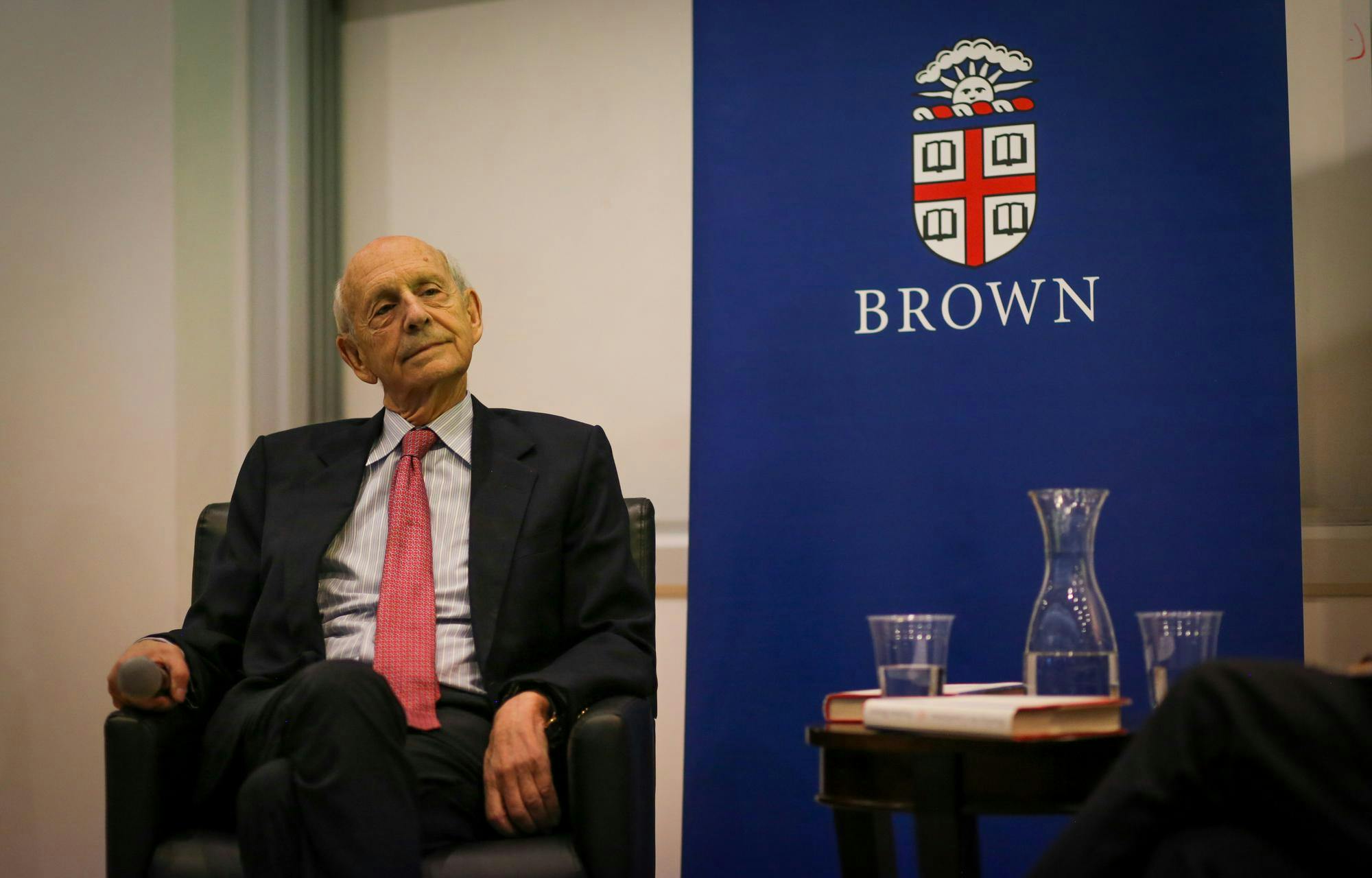 An older gentleman wearing a black suit and a red tie sits in a chair next to a blue banner inscribed with Brown's logo.