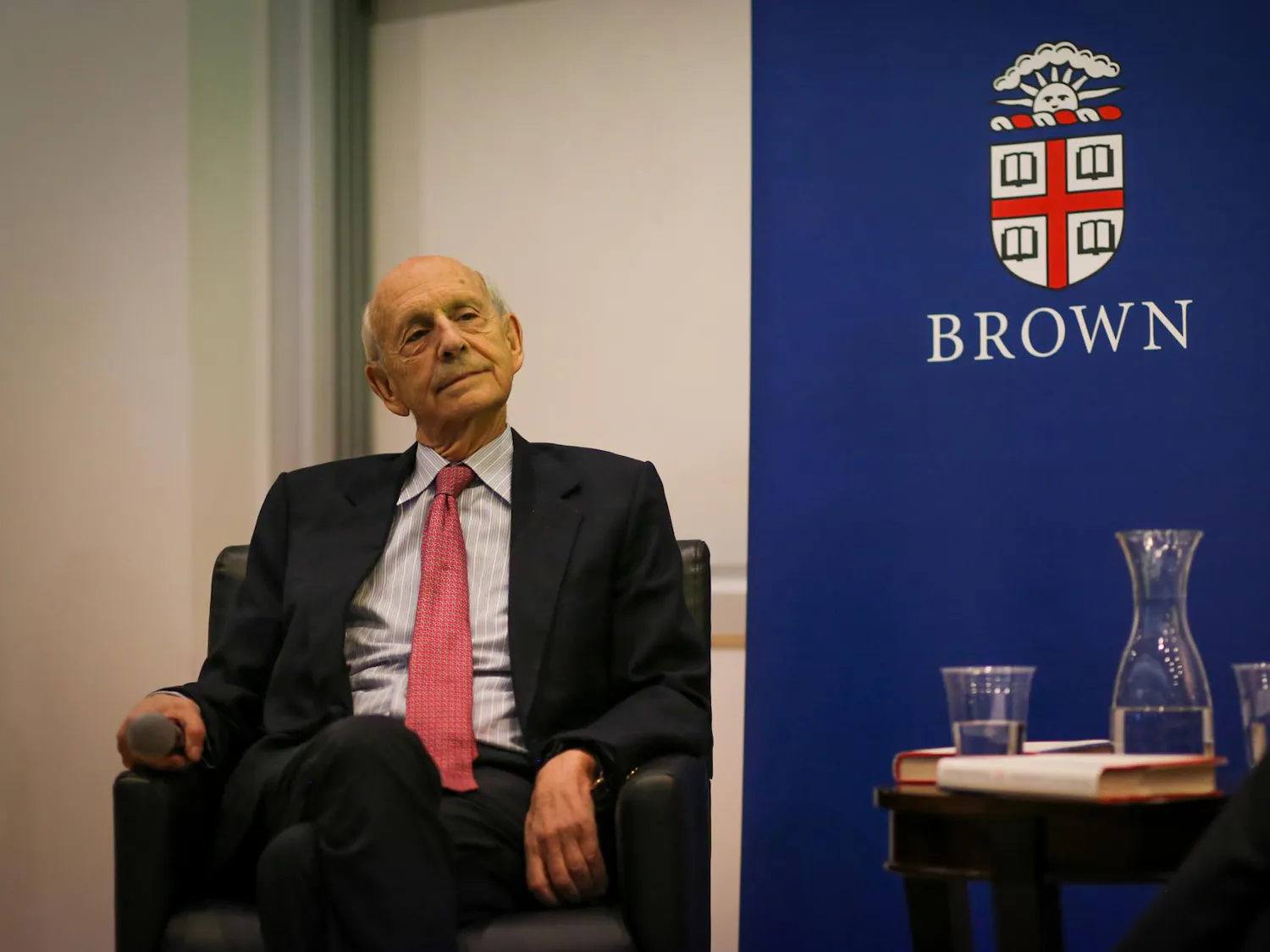 An older gentleman wearing a black suit and a red tie sits in a chair next to a blue banner inscribed with Brown's logo.