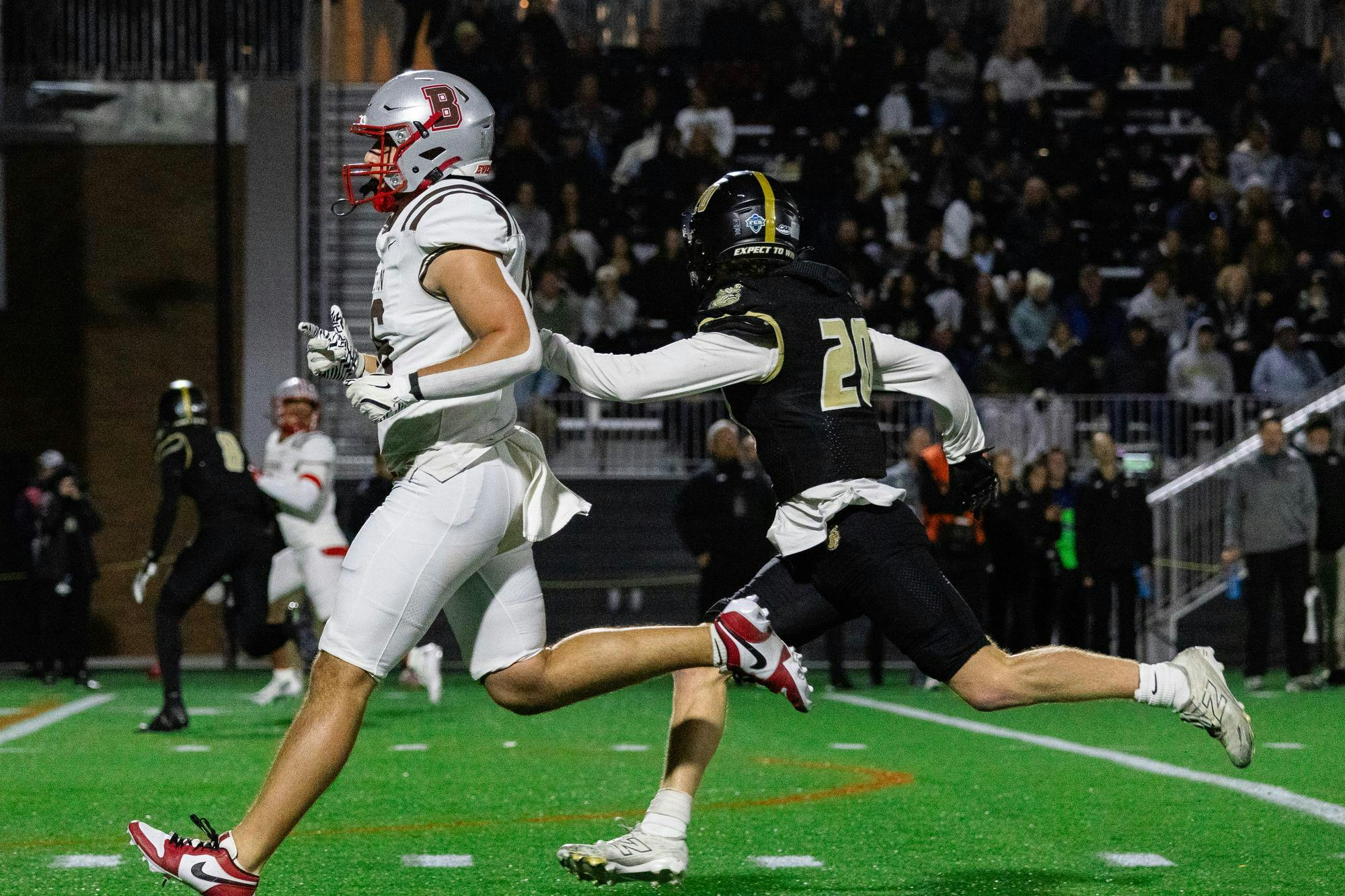 Photo of a Brown football player followed by a Bryant football player running down the field viewed from the side.